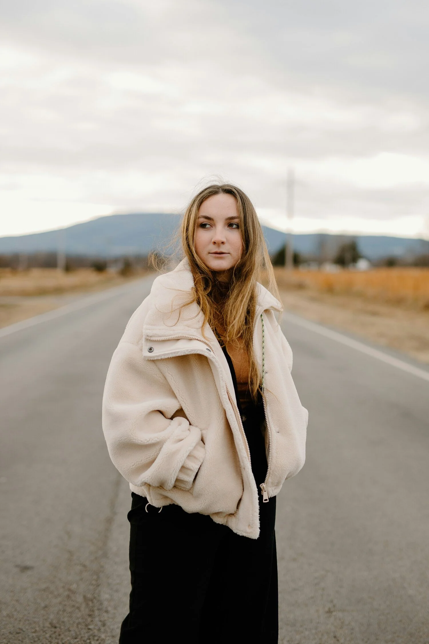 Young woman with light brown hair wearing a beige fleece jacket and black pants standing on an empty rural road with mountains in the background under gray cloudy sky.