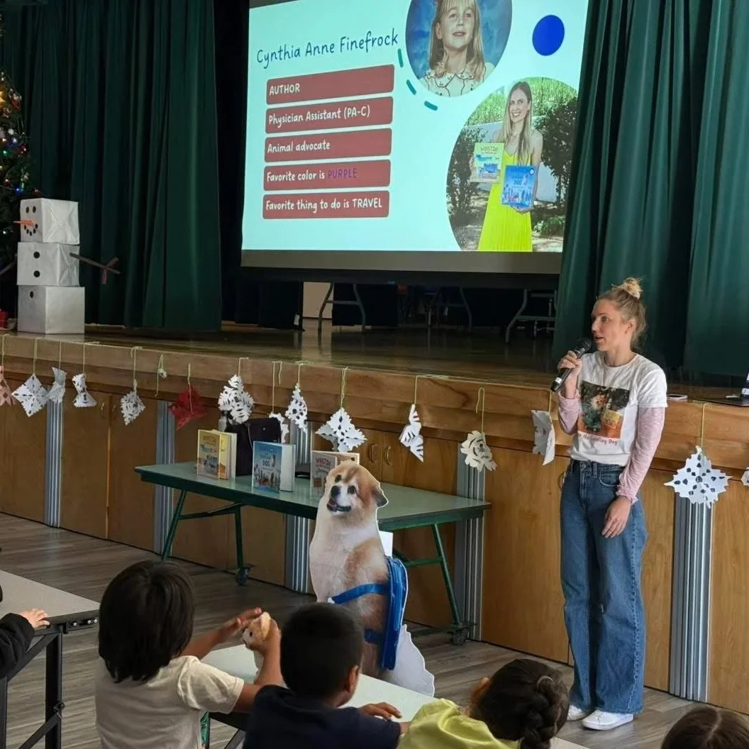 A classroom presentation with children seated at tables, a woman holding a microphone standing in front of the audience, decorated with a holiday tree and paper snowflakes, and a large screen displaying information about named Cynthia Anne Finefrock,