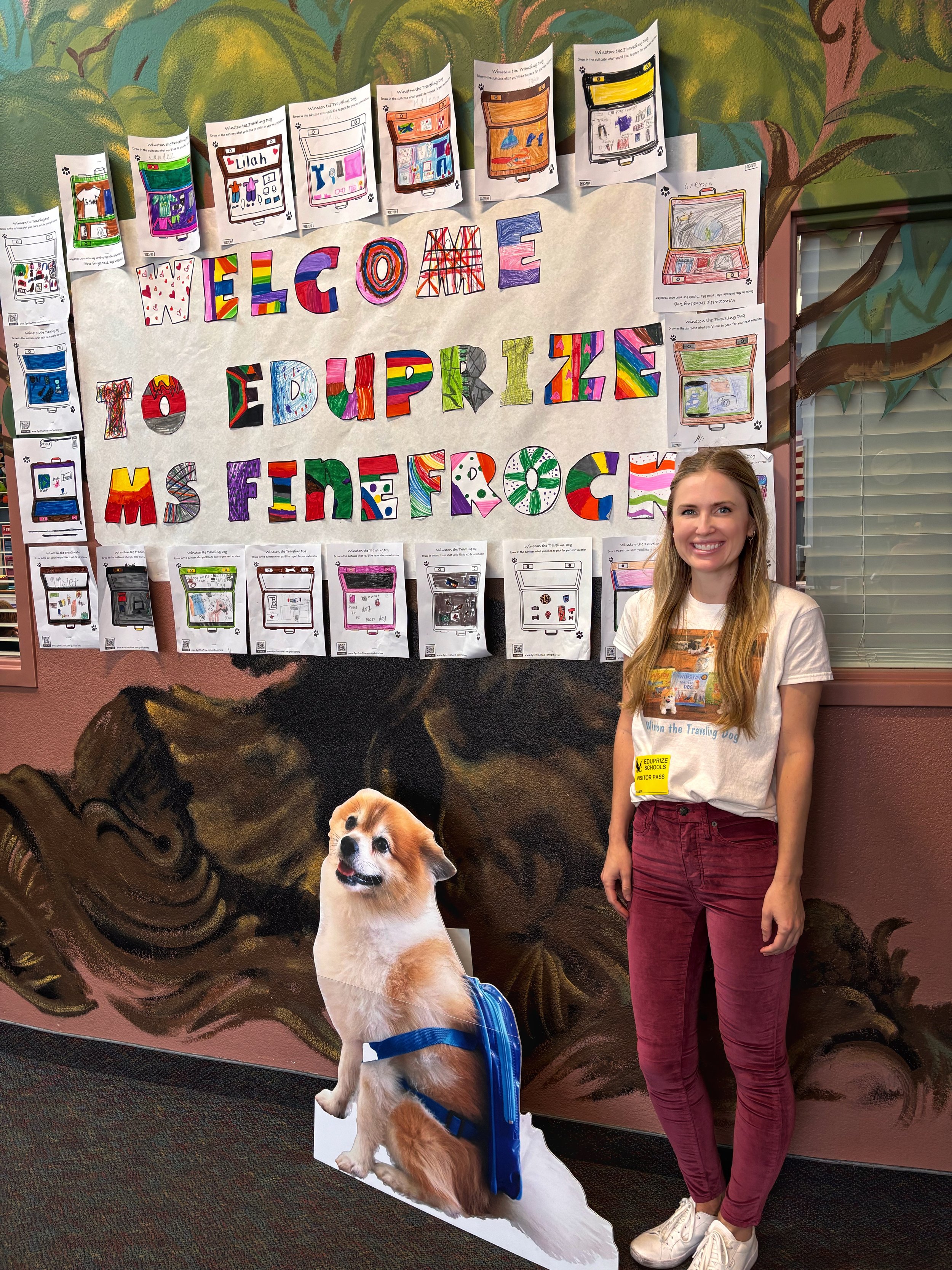 Cynthia Finefrock is standing in front of a colorful classroom bulletin board that reads "Welcome to EduPrize MS FIMEFROCK" with various student artwork and drawings, and a large cutout of a dog with a backpack is on the floor next to her.