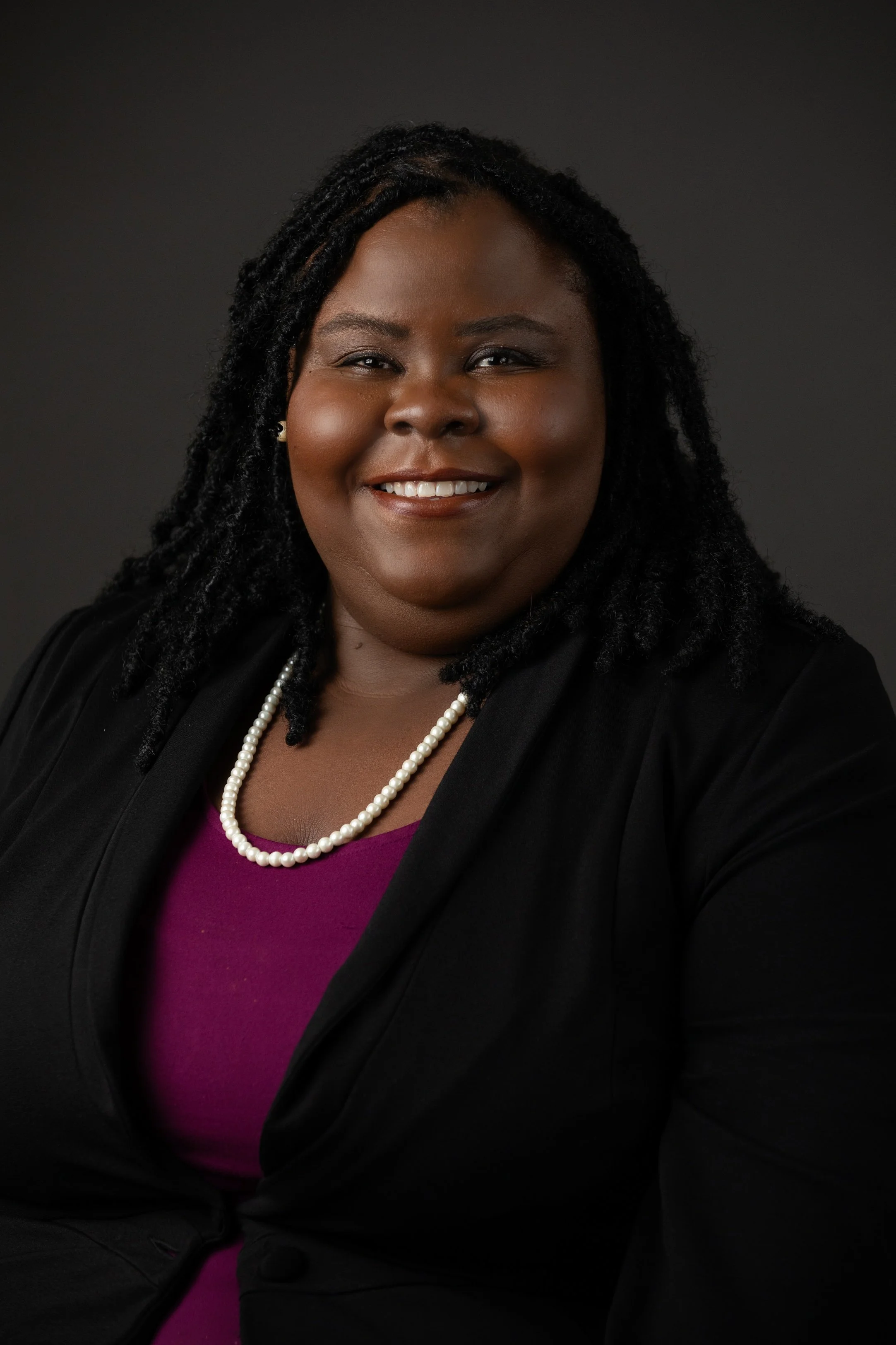 A professional portrait of a smiling Black woman with shoulder-length curly hair, wearing a black blazer, a purple top, and a pearl necklace against a dark background.