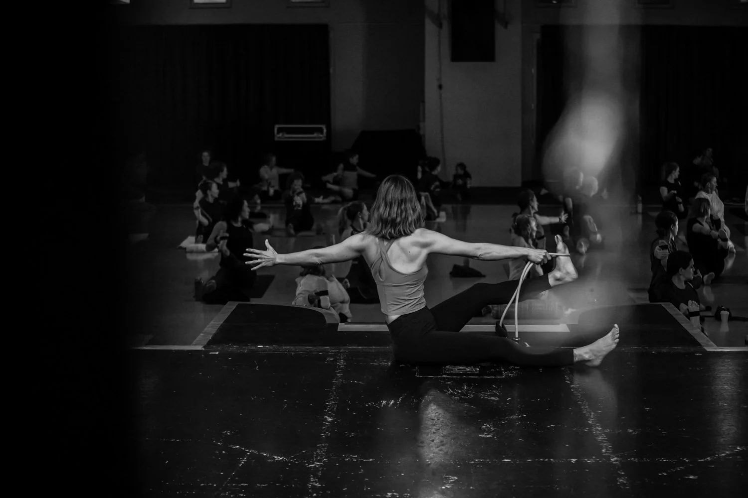 Black and white photo of a dance instructor demonstrating a split pose on stage with her arms outstretched, while a group of children practice seated on the floor in a dance studio.