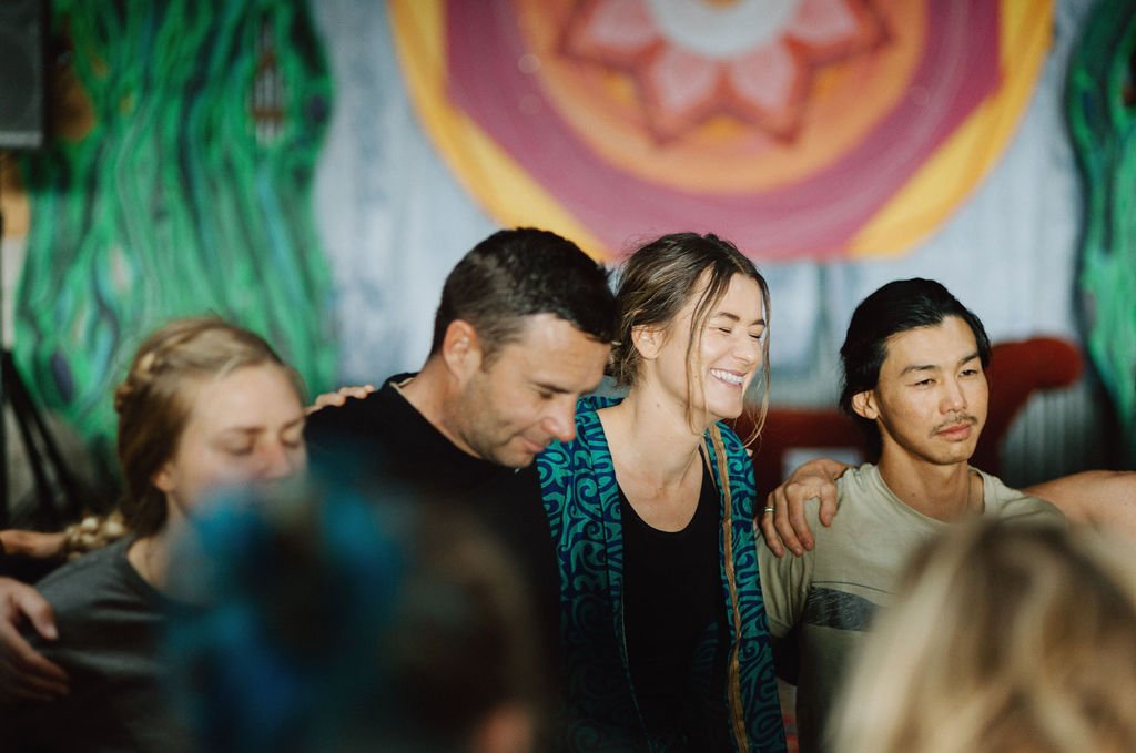 Group of people with their eyes closed, embracing, and smiling during a gathering or ceremony with colorful background artwork.