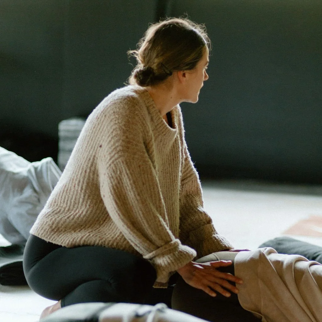 A woman sitting on her knees with a beige sweater, looking off to the side. She appears to be in a calm, indoor setting.