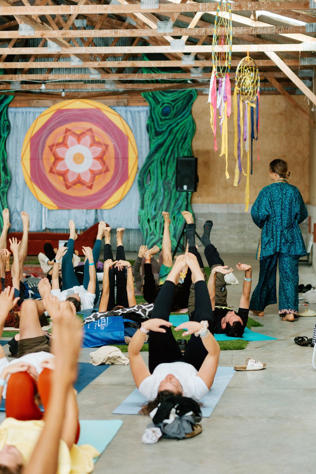 People participating in a yoga class in a rustic indoor space, with colorful dreamcatchers hanging from the ceiling and a large decorative mandala-style wall hanging in the background.
