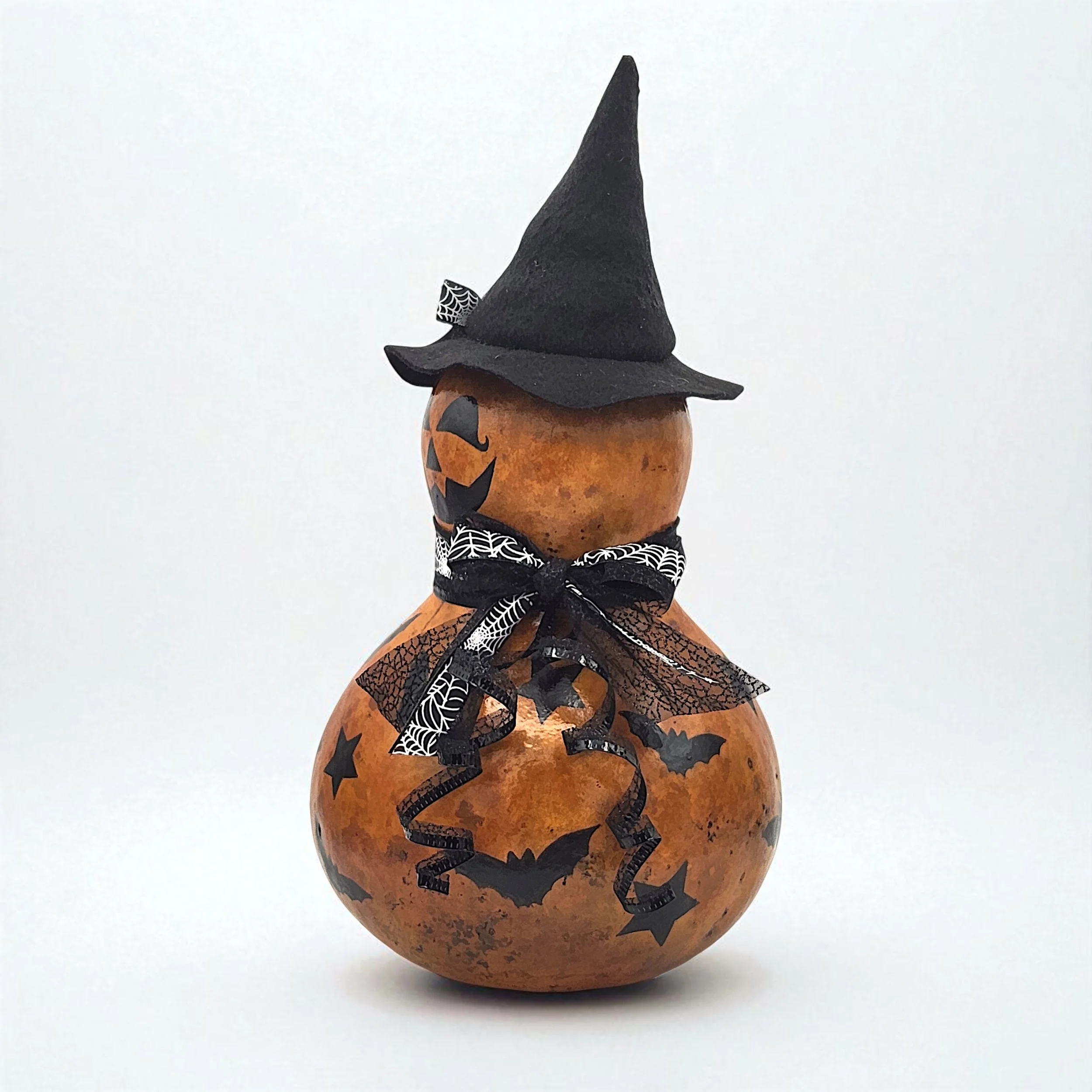 Side profile of jack-o-lantern bottle gourd featuring a painted face, layered stencil details, and a fancy ribbon bow at the neck, with the witch hat tilted playfully to one side.