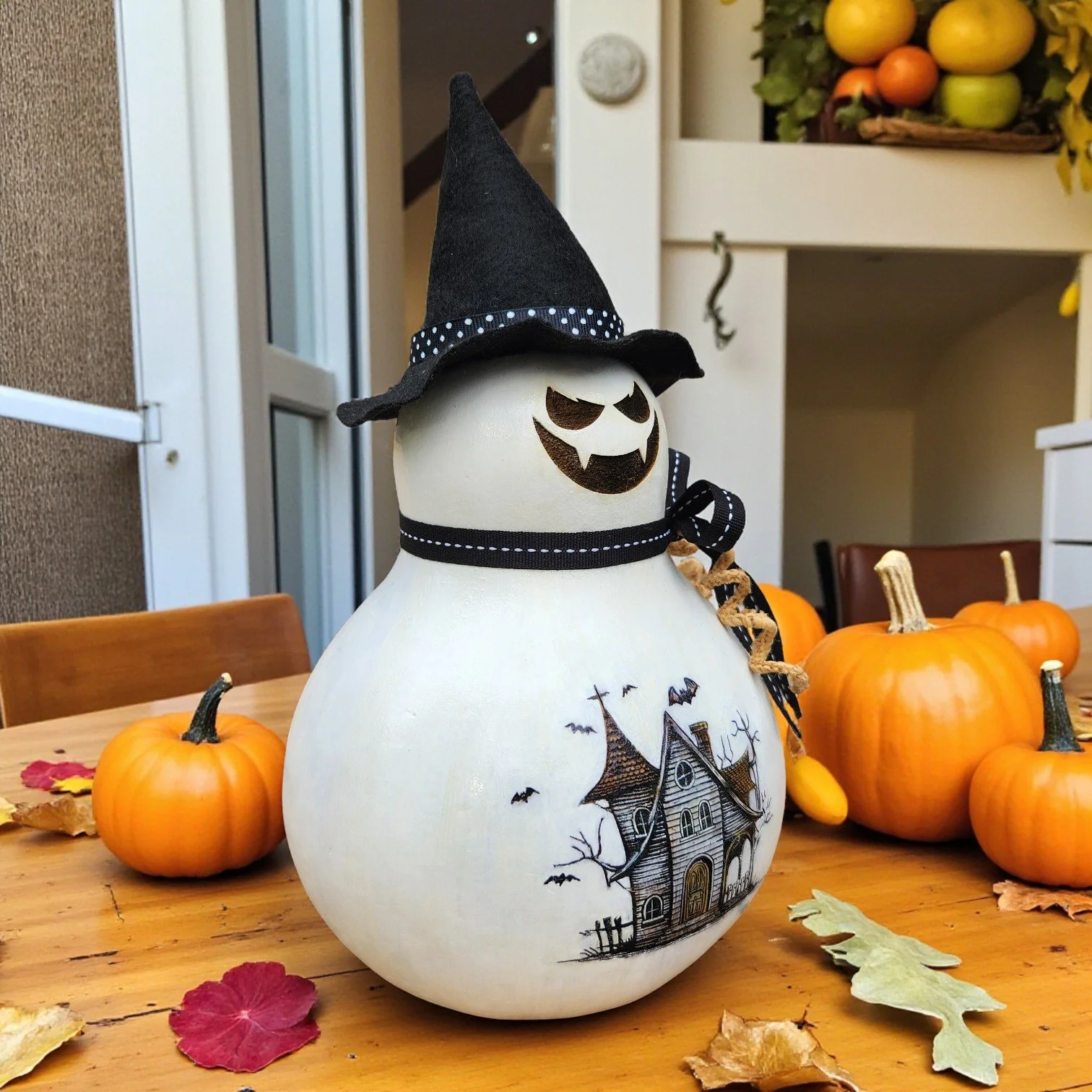 Hand-painted bottle gourd showing both spooky face and haunted house engravings, with layered black, white, and gold ribbon.