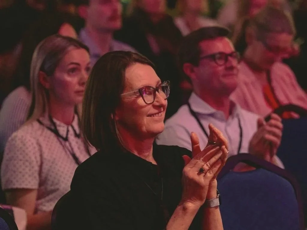 An audience of adults seated and watching a presentation, with a woman in the foreground clapping and smiling.