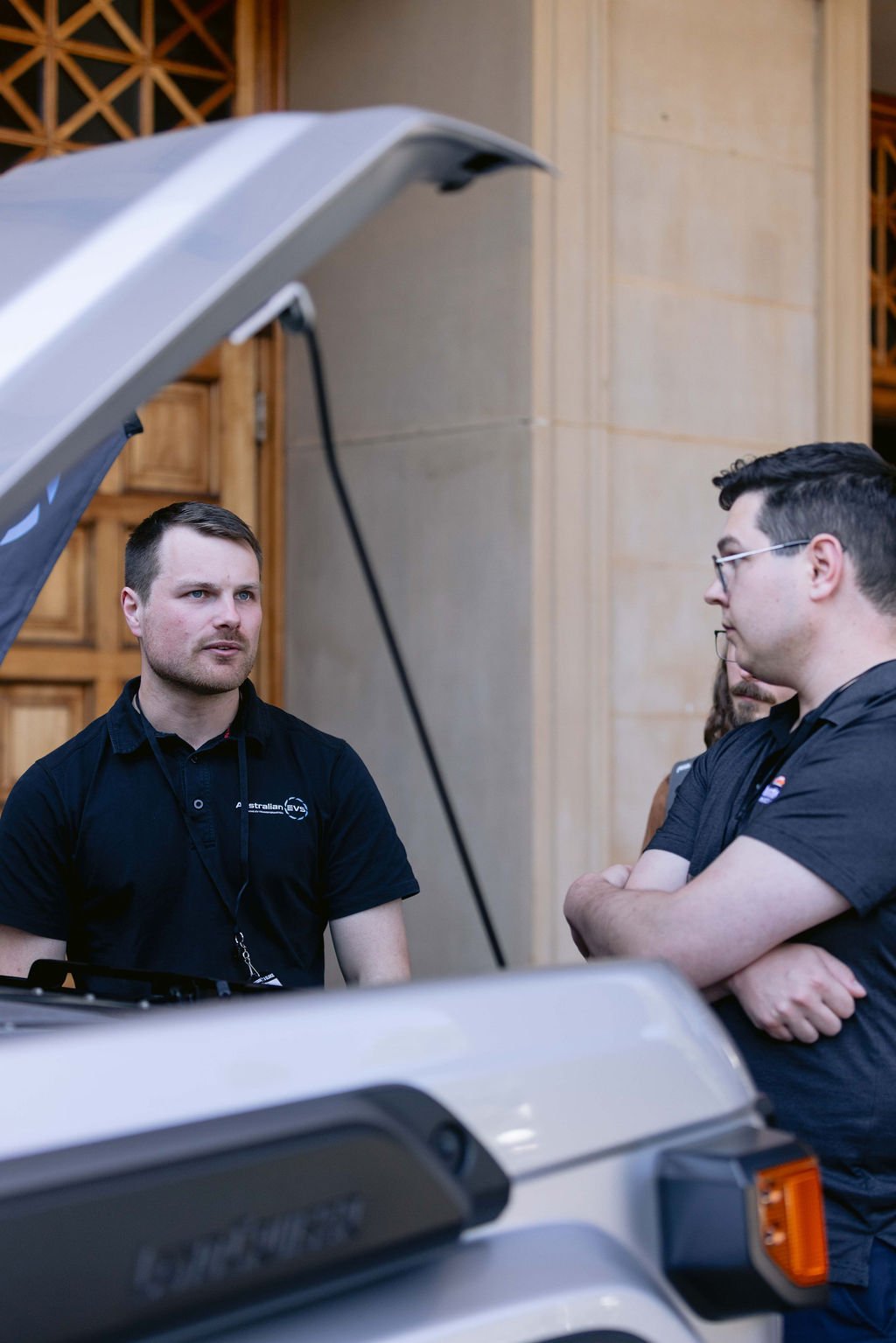 Two men in black shirts having a conversation outdoors, with a vehicle hood open nearby.