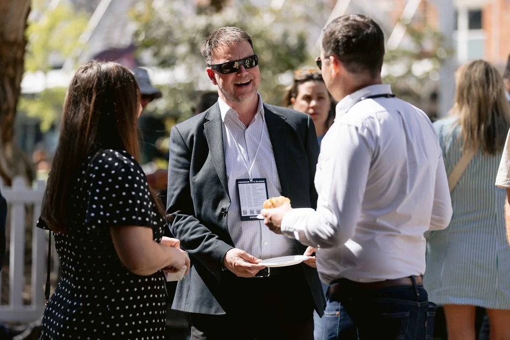 Three people conversing outdoors at a daytime event; a man in sunglasses and a suit talking to a man in a white shirt, with a woman in a polka dot dress nearby, and other people blurred in the background.