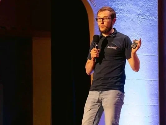 Man speaking on stage at a conference or event, holding a microphone in one hand and a small device in the other, with a blue-lit wall in the background.