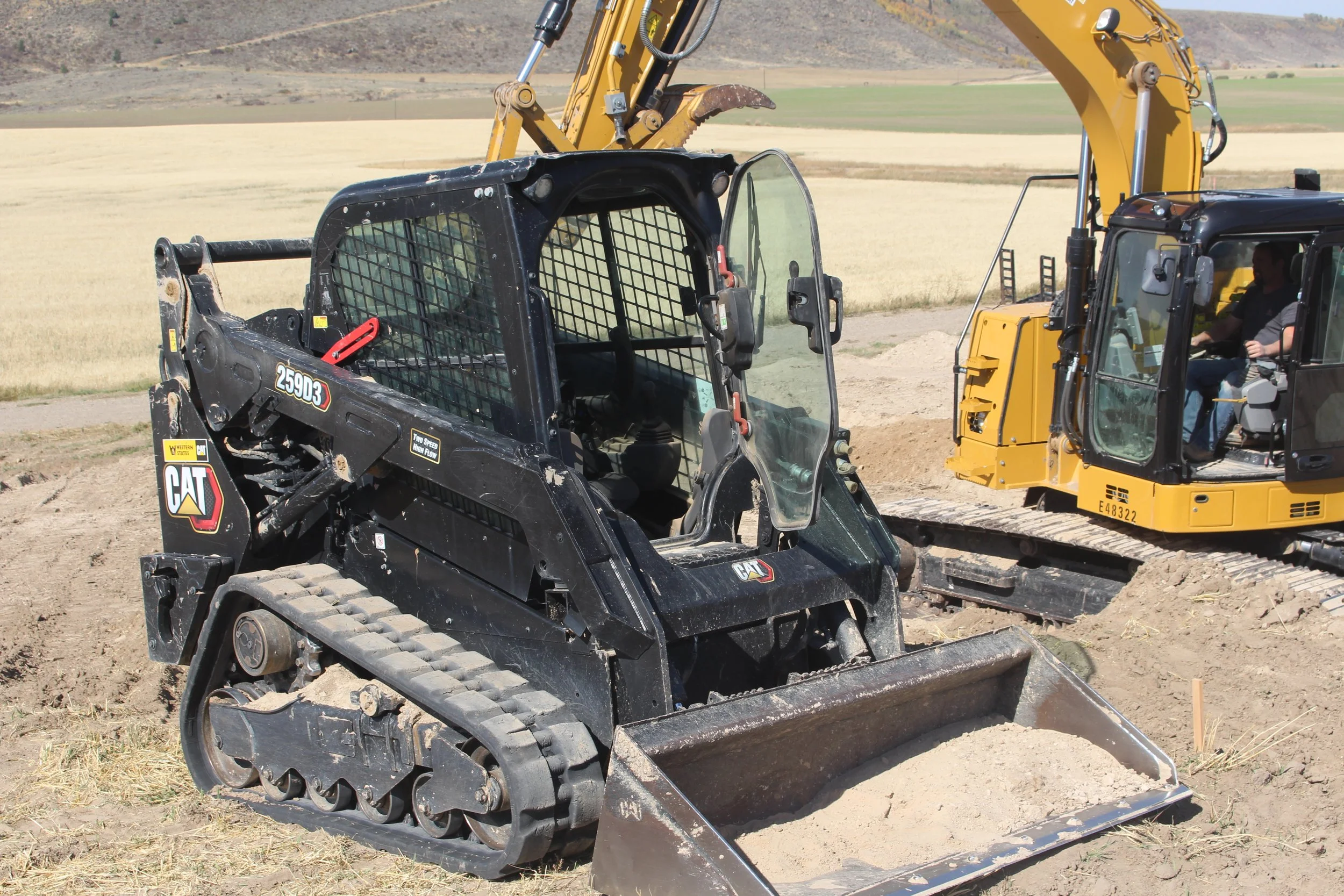 A black CAT compact track loader with a bucket attachment, working on a construction site with a larger yellow excavator nearby. The site is in a rural area with open fields and mountains in the background.