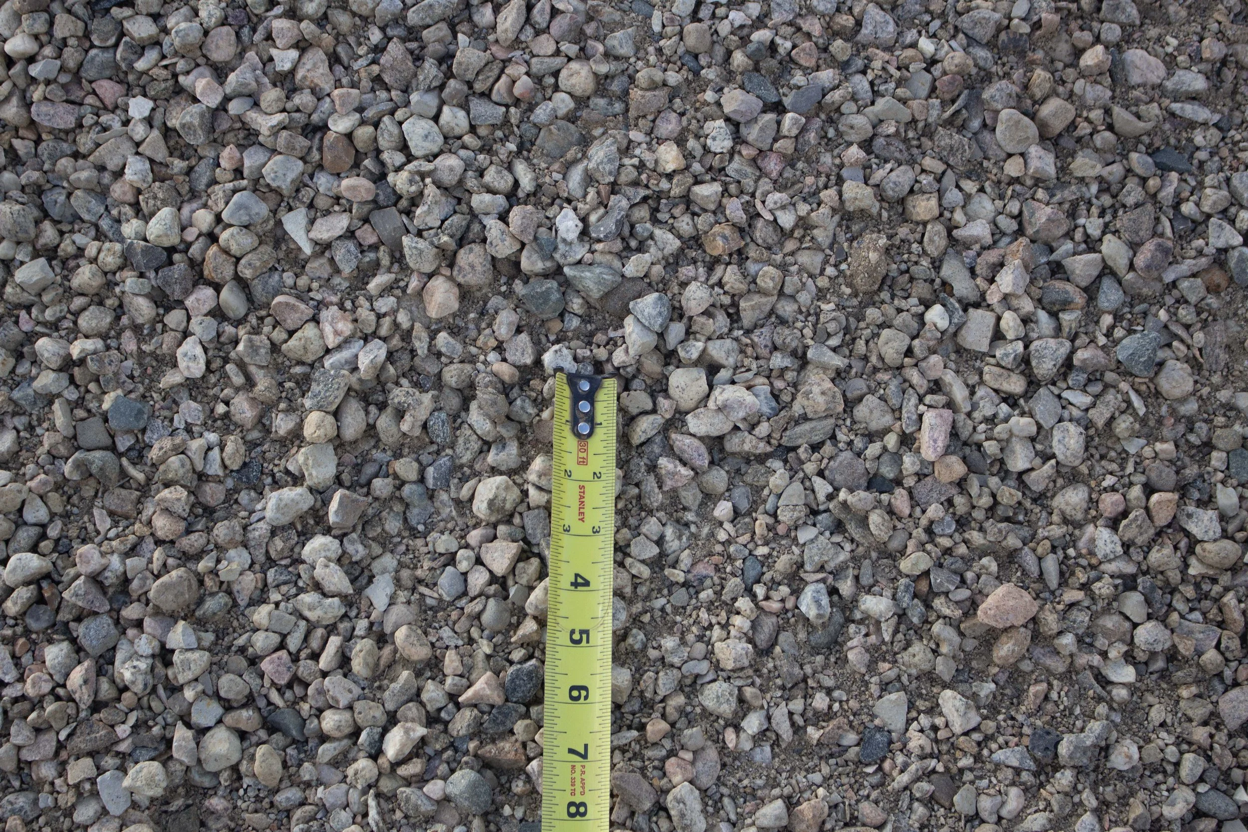 Close-up of gravel with a yellow measuring tape showing approximately 6 inches.