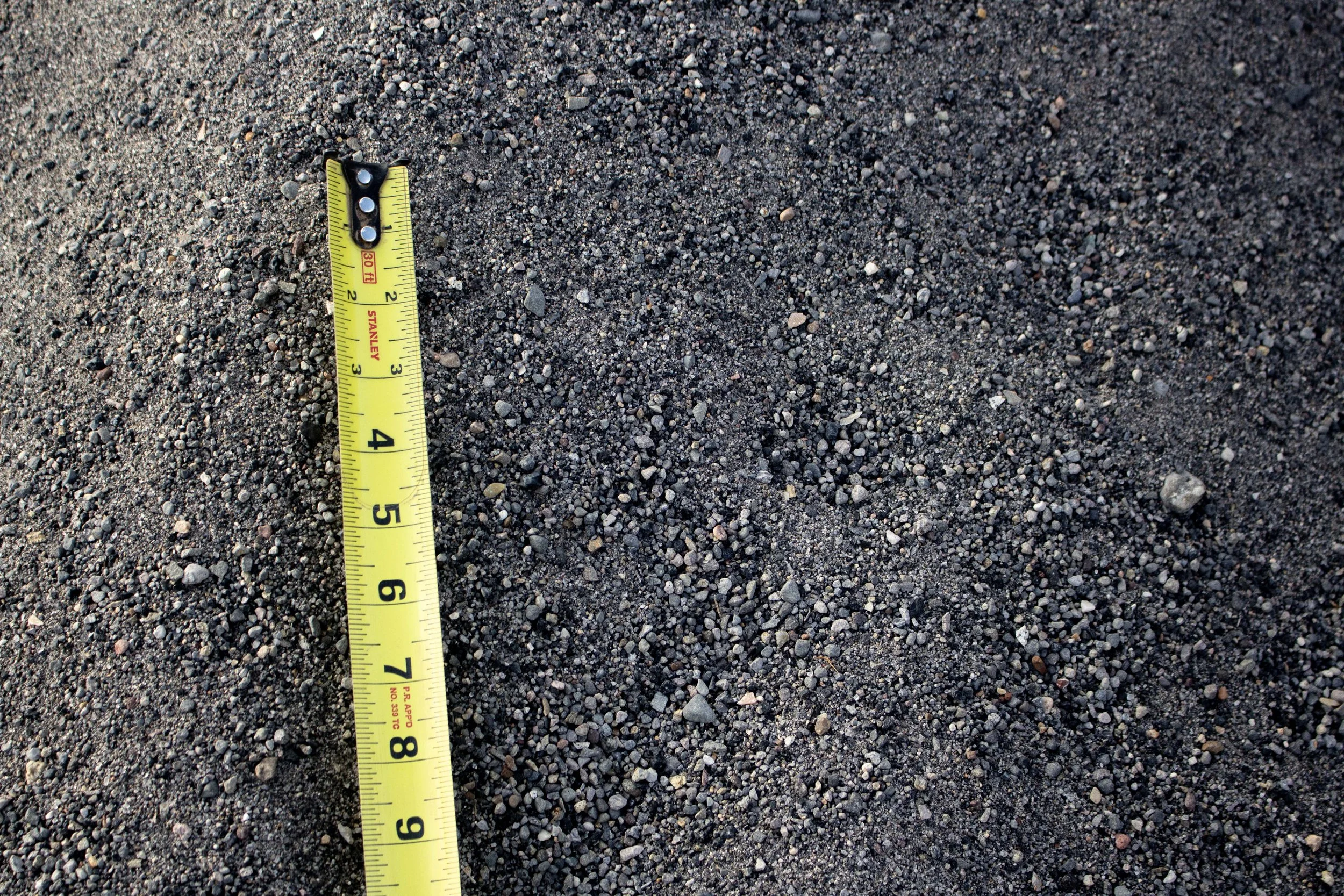 Close-up of a yellow measuring tape on dark, gritty gravel or sand surface.