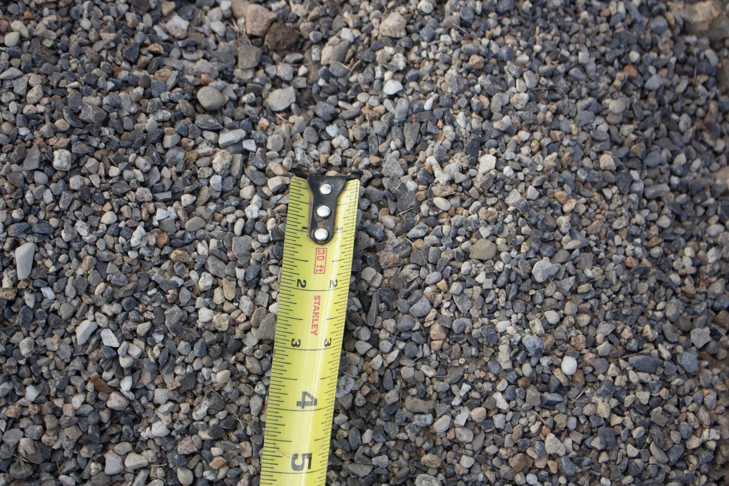 A yellow tape measure on a bed of small rocks and gravel, measuring approximately 3 inches.