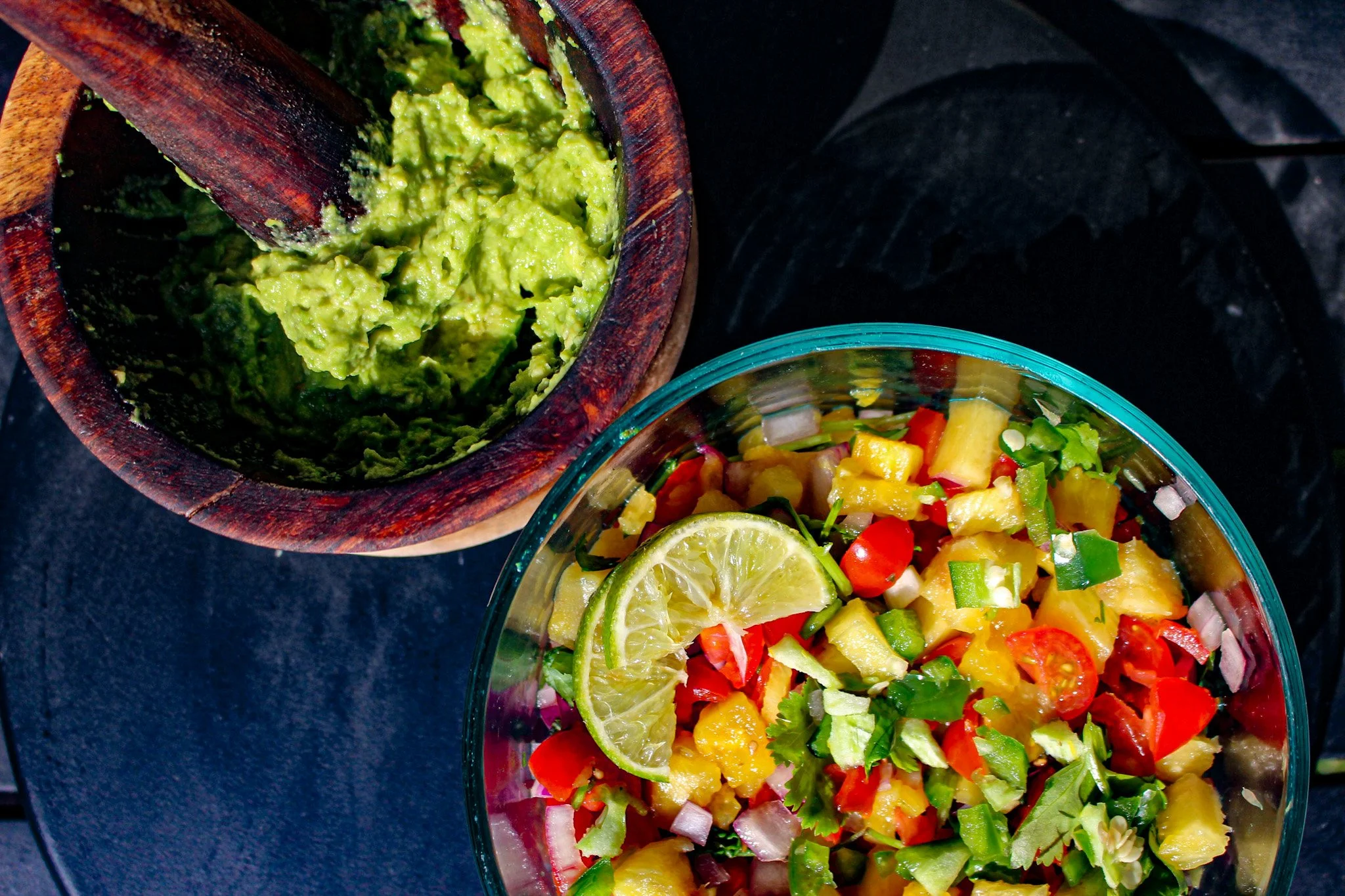 A bowl of colorful diced fruits and vegetables garnished with a lime wedge and chopped herbs, next to a wooden bowl of mashed avocado.