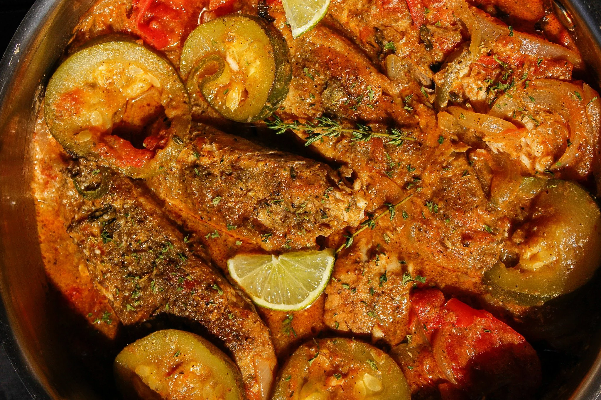 Close-up of cooked fish fillets in a tomato-based sauce with sliced jalapeños and a lemon wedge, garnished with herbs.