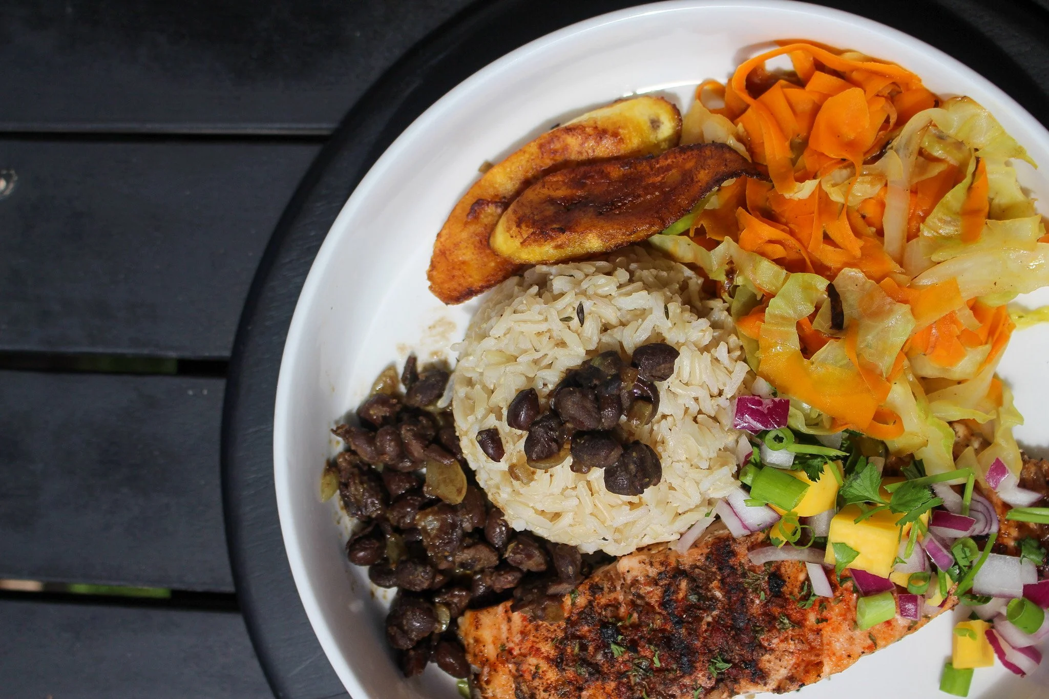 Plate with grilled fish, black beans, rice with black beans, fried plantain, and colorful salad with lettuce, tomato, red onion, and chopped yellow pepper.