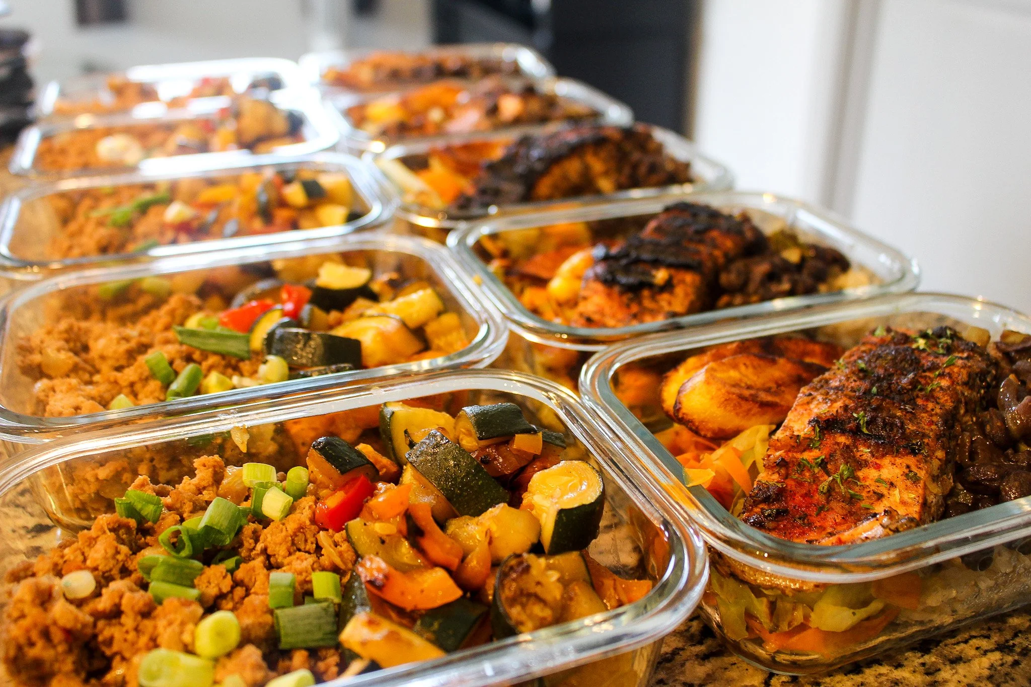 Multiple glass meal containers filled with cooked vegetables, ground meat, and roasted chicken on a kitchen countertop.