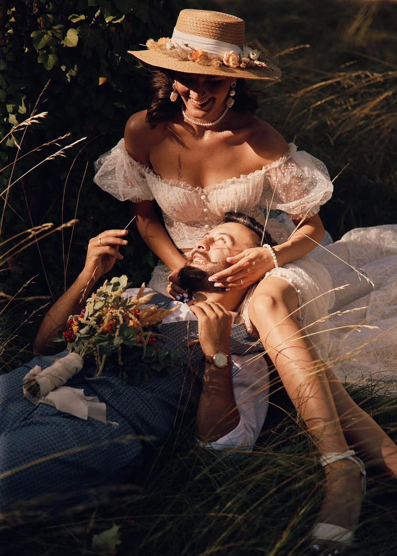Bride sitting with a bouquet of flowers on the ground, groom next to her