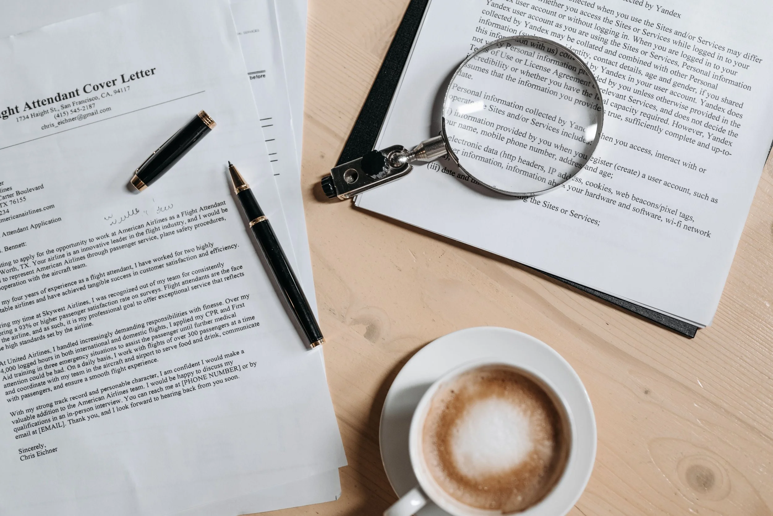 Desk with various papers, a magnifying glass, a pen, and two cups of coffee on a wooden surface.
