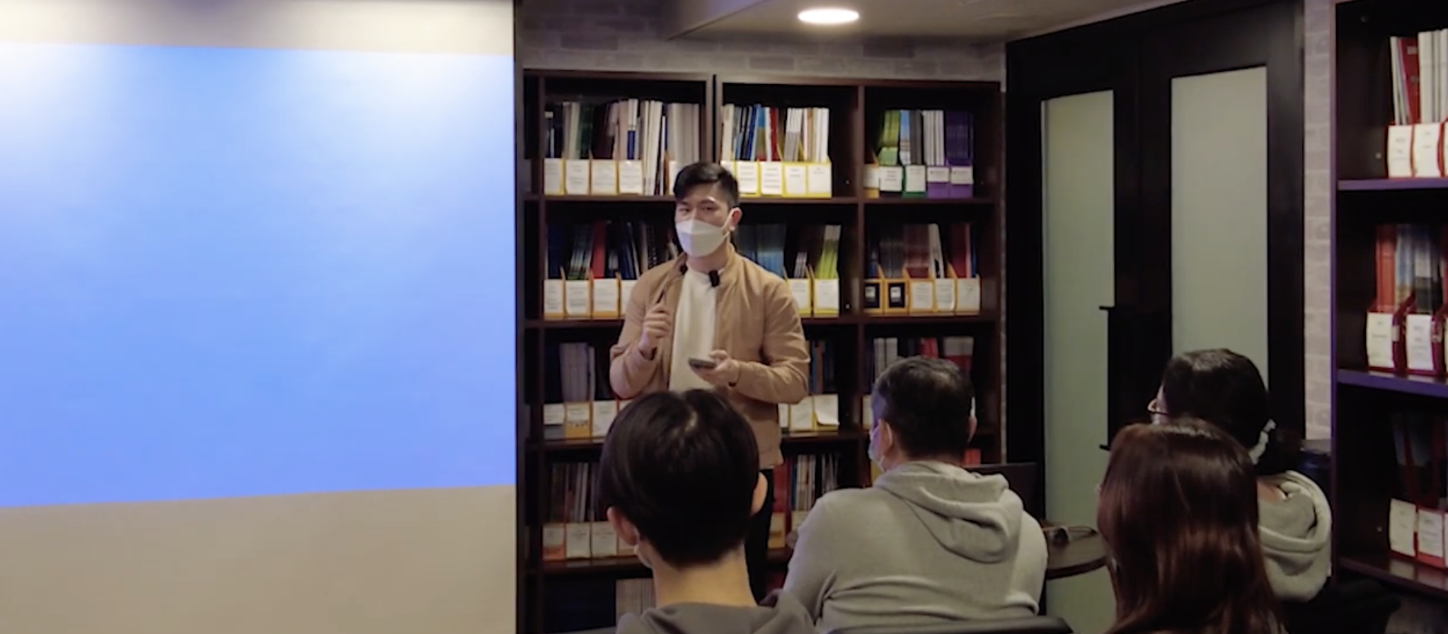 A group of people sitting in a room listening to a speaker wearing a beige jacket and a face mask. The speaker is standing in front of bookshelves filled with books and holding a mobile phone. There is a blue screen or wall on the left side of the image.