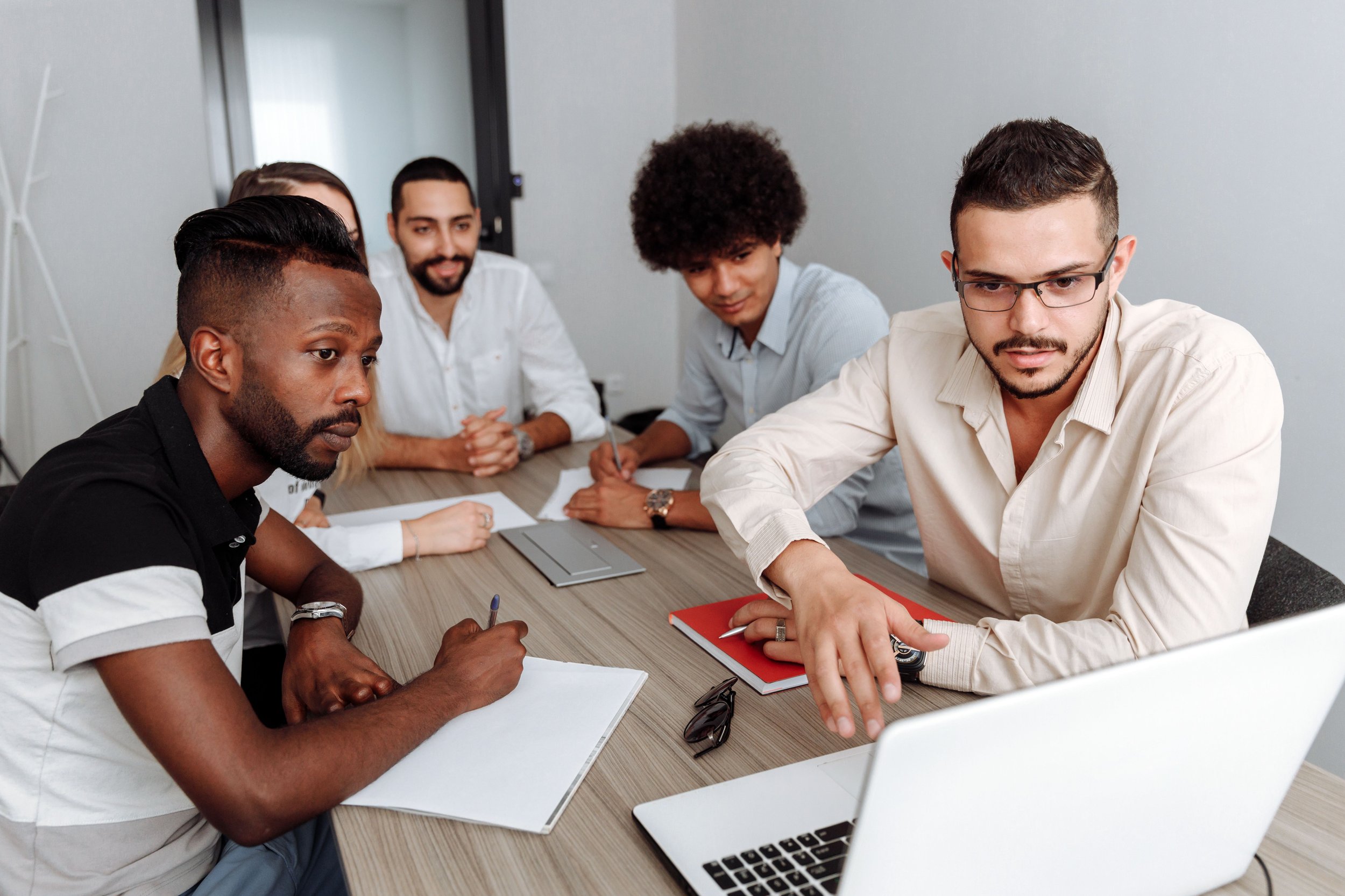 A diverse group of five young adults sitting around a wooden table in a modern office space, engaged in a discussion or meeting, with notebooks, pens, and a laptop visible on the table.