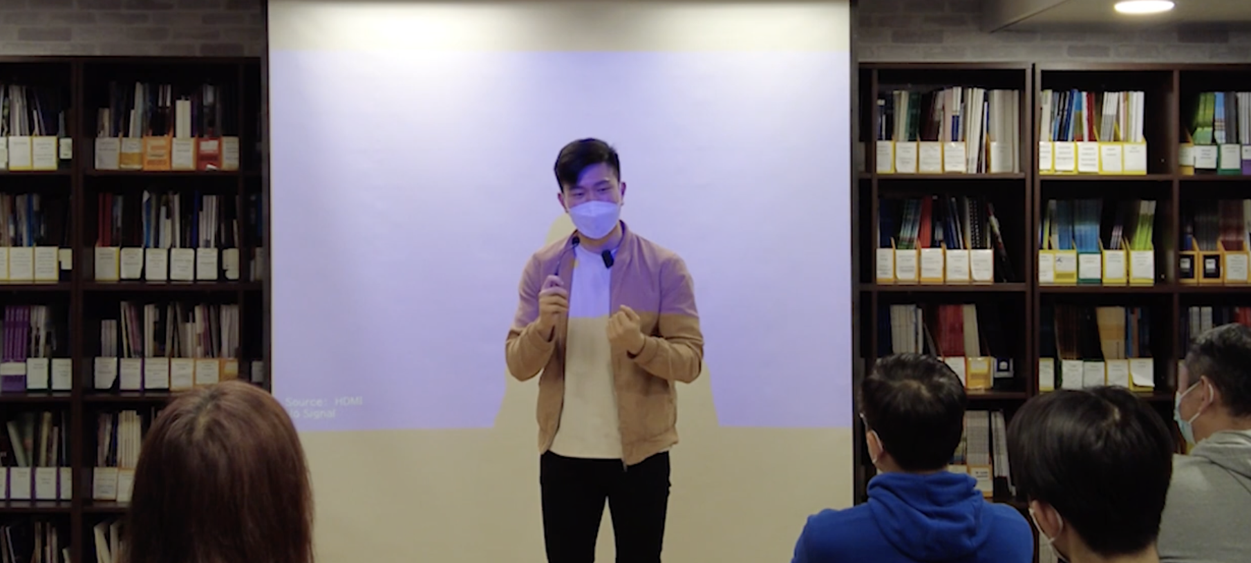 Young man wearing a face mask giving a presentation in front of a white screen in a library, with bookshelves on either side and an audience wearing face masks facing him.