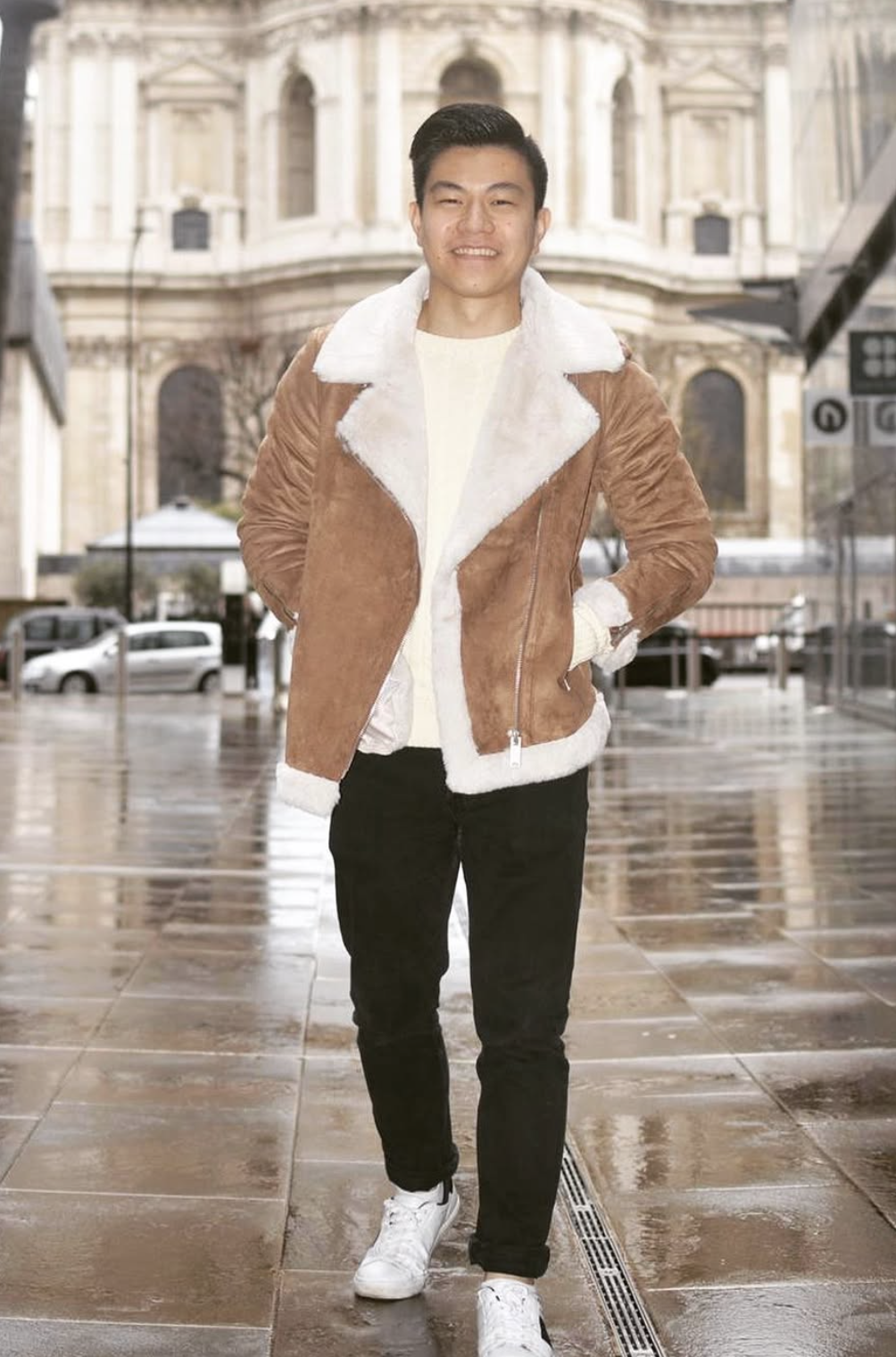 A young man smiling and walking outdoors on a wet, reflective pavement in front of a historic building, wearing a brown shearling-lined jacket, white sweater, black pants, and white sneakers.