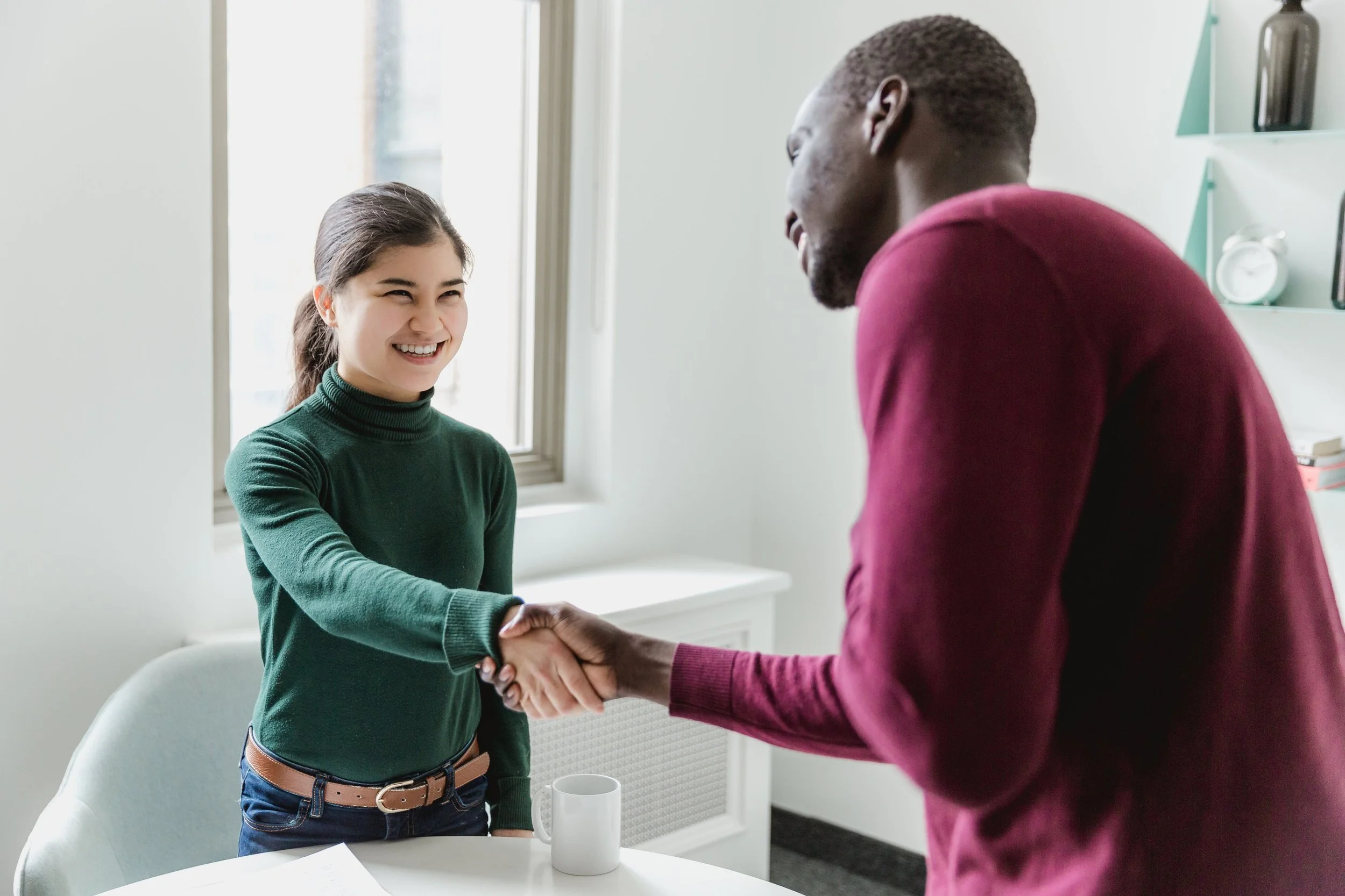 A woman with dark hair in a ponytail, wearing a green turtleneck, smiling and shaking hands with a man in a red sweater in a bright office with a window and shelves in the background.