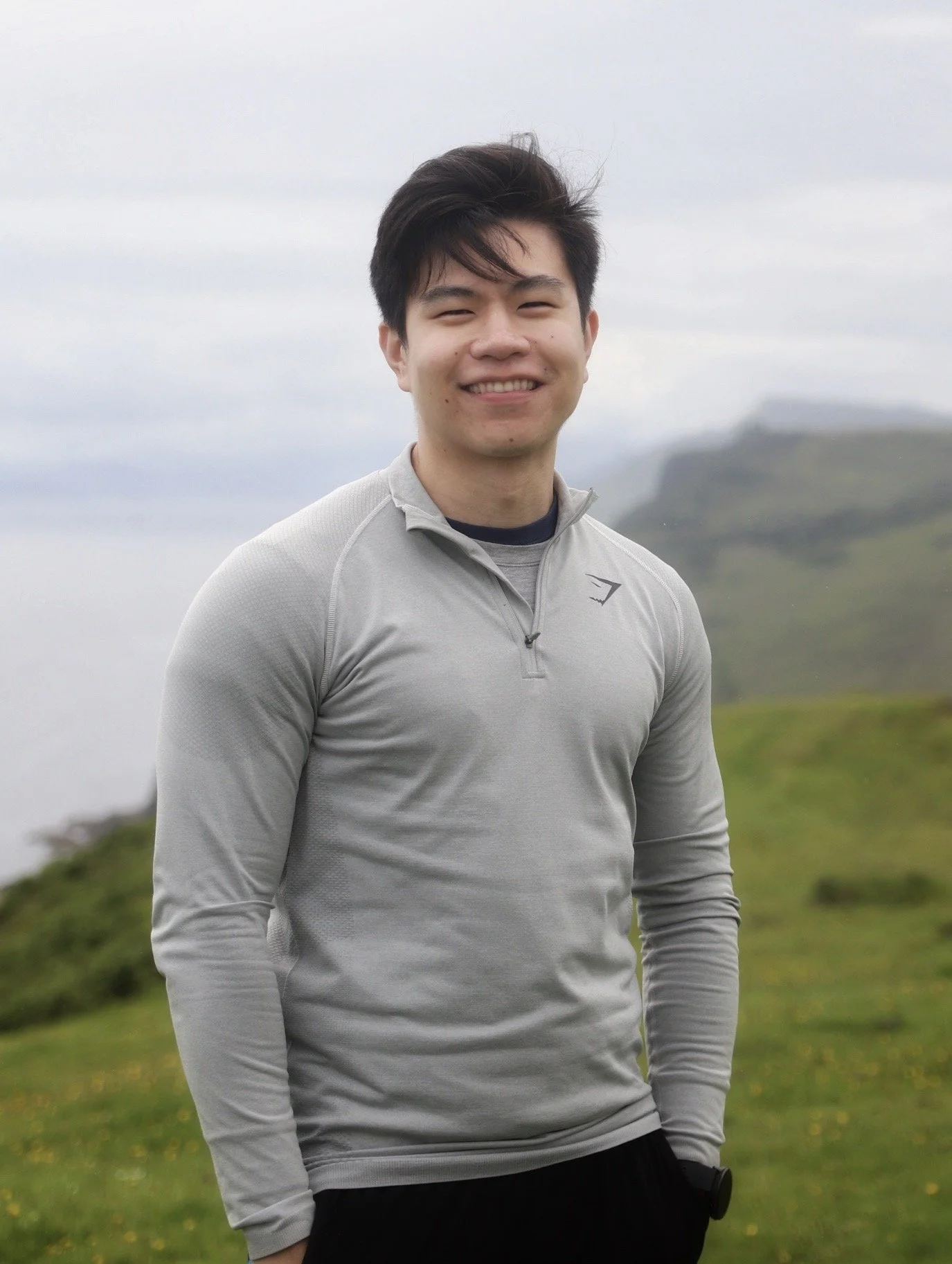 A young man smiling outdoors on a cloudy day, wearing a light gray zip-up athletic jacket with a black shirt underneath, standing in front of a grassy landscape with hills in the background.
