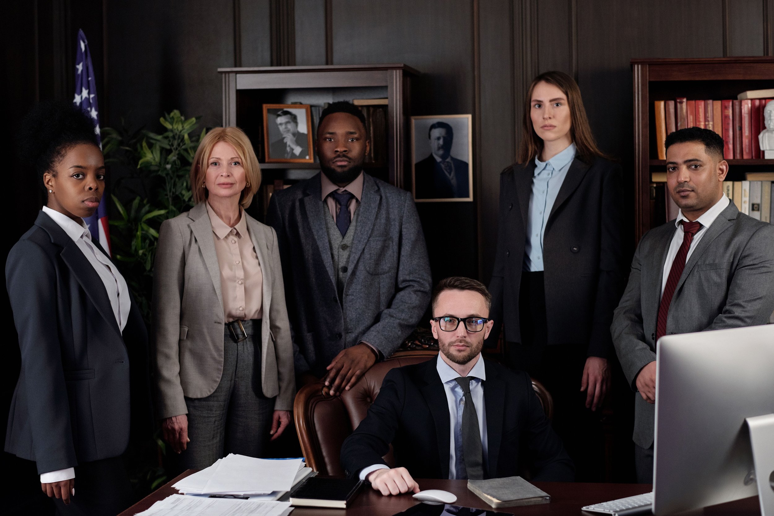 A group of six diverse professionals in formal attire in a dark office. Four standing behind a seated man at a desk with papers, a computer, and a mouse. Background includes a bookshelf, framed photos, a flag, and a potted plant.