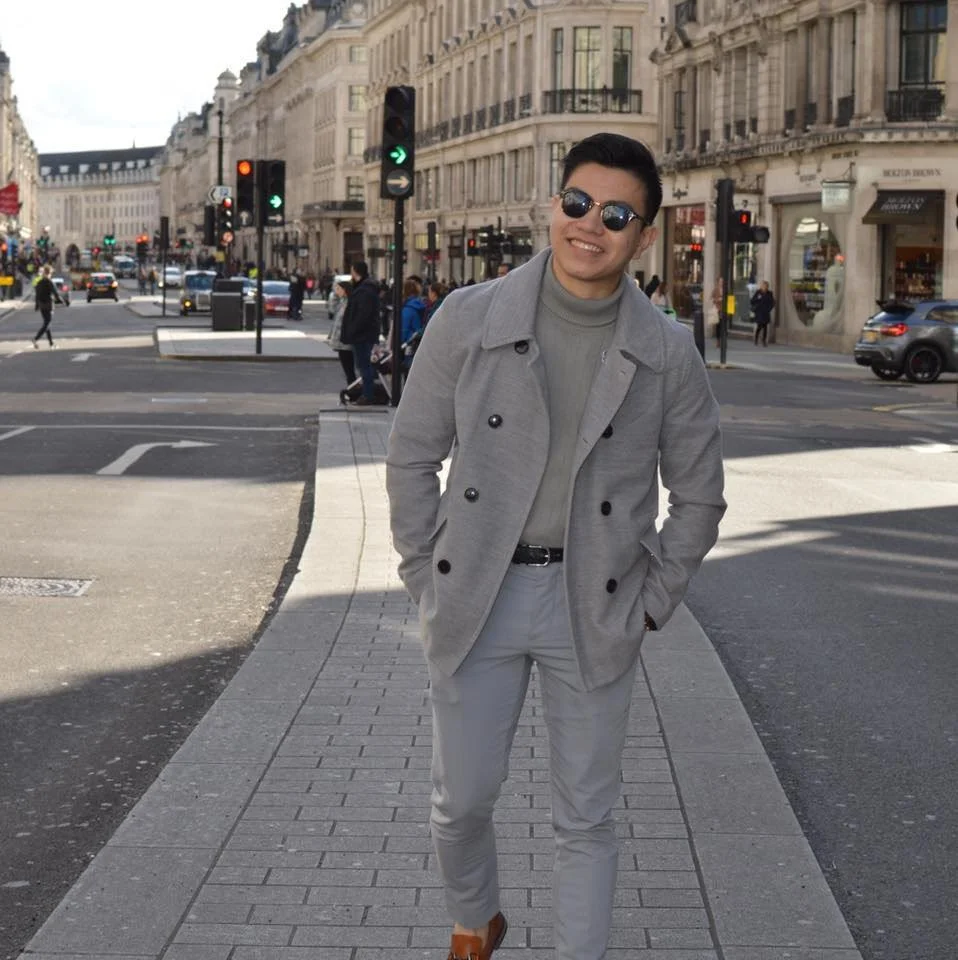 Young man in sunglasses smiling at camera, dressed in gray coat and pants, walking on city sidewalk with historic buildings and busy street in the background.