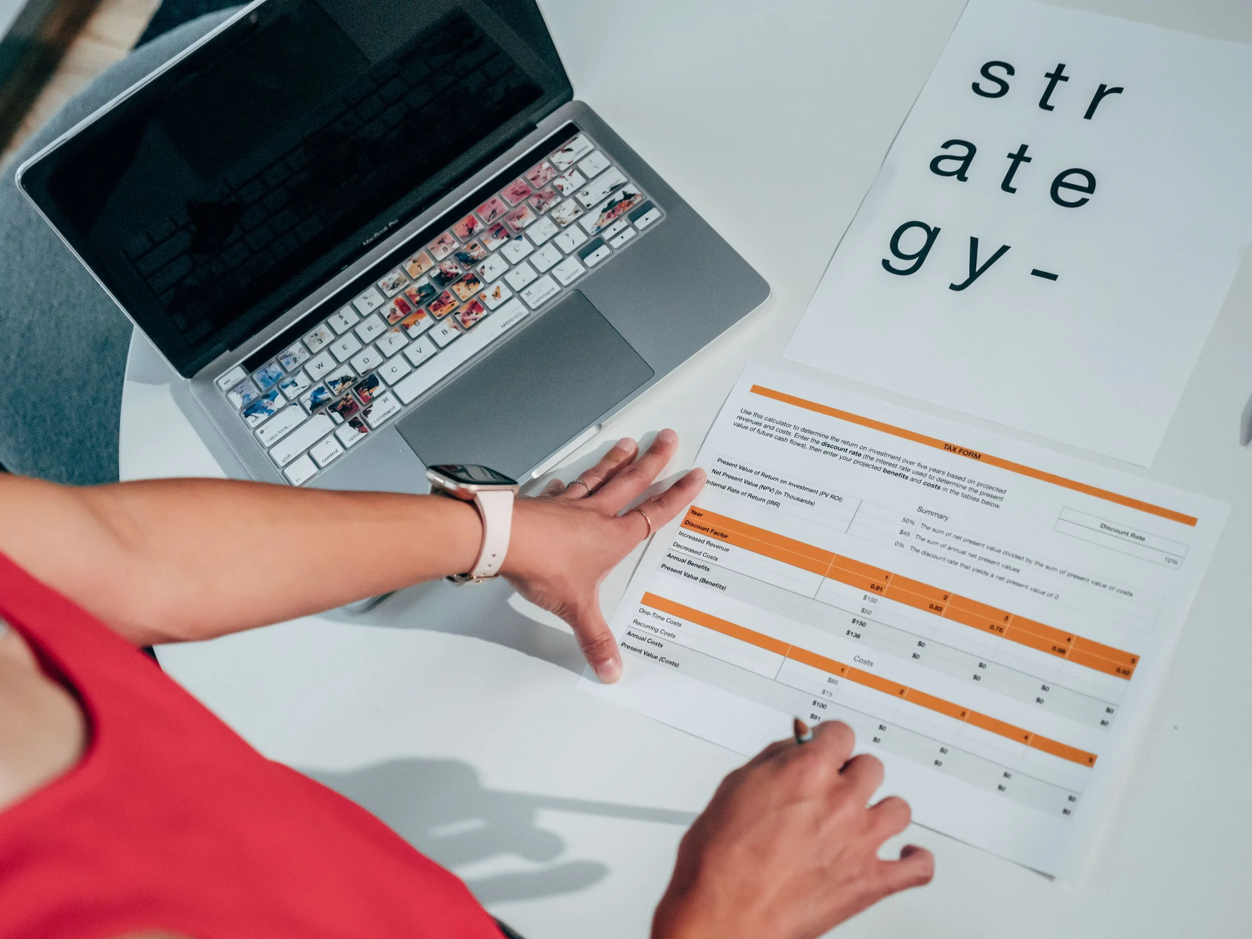 Person in red shirt pointing at a financial spreadsheet on a white table, with a silver laptop nearby and a sign saying 'strate gy-'.