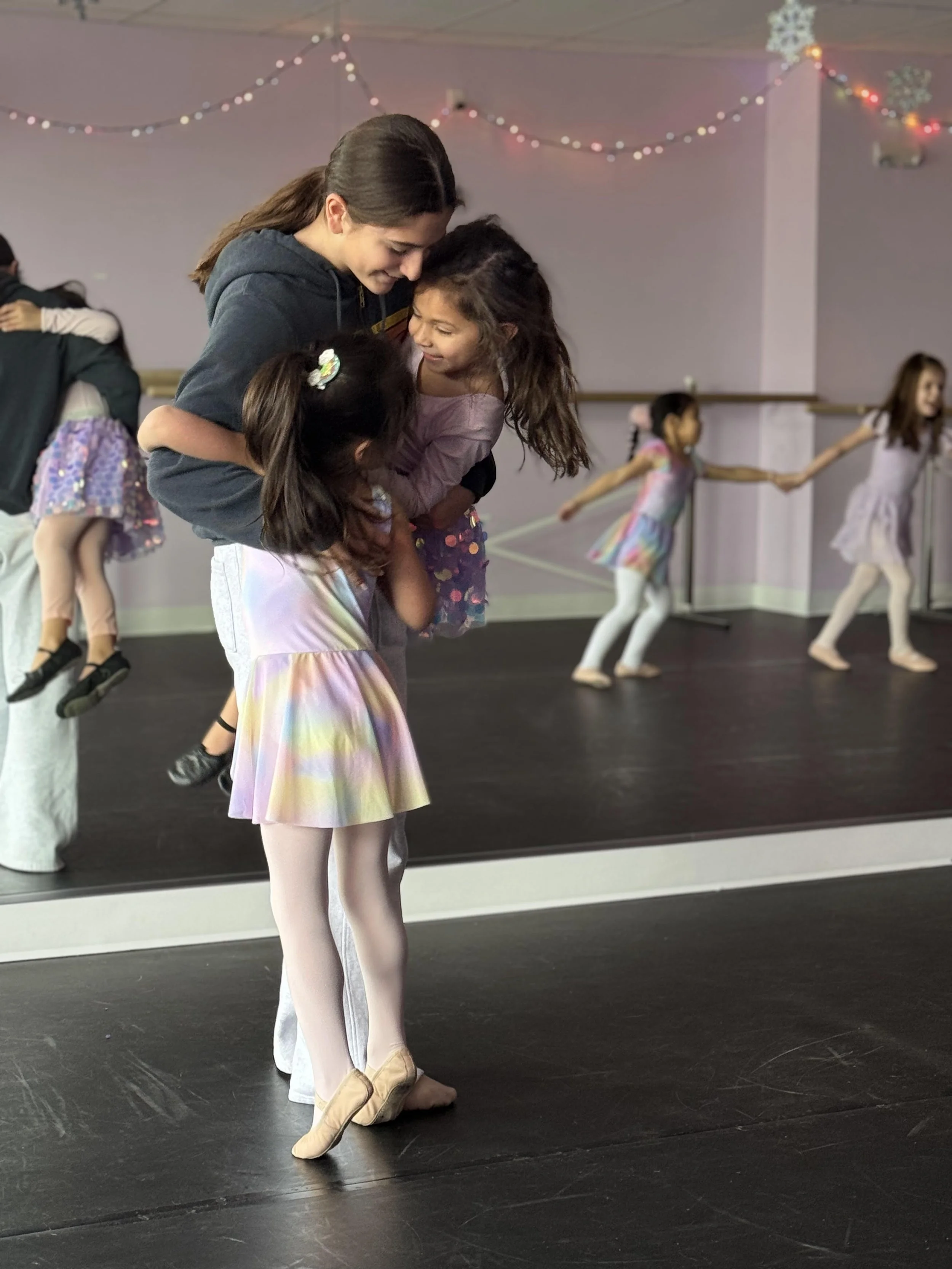 A woman and three young girls at a ballet studio, with the woman lifting one girl in the air while the other girls practice ballet in the background. Holiday lights and snowflake decorations hang overhead.