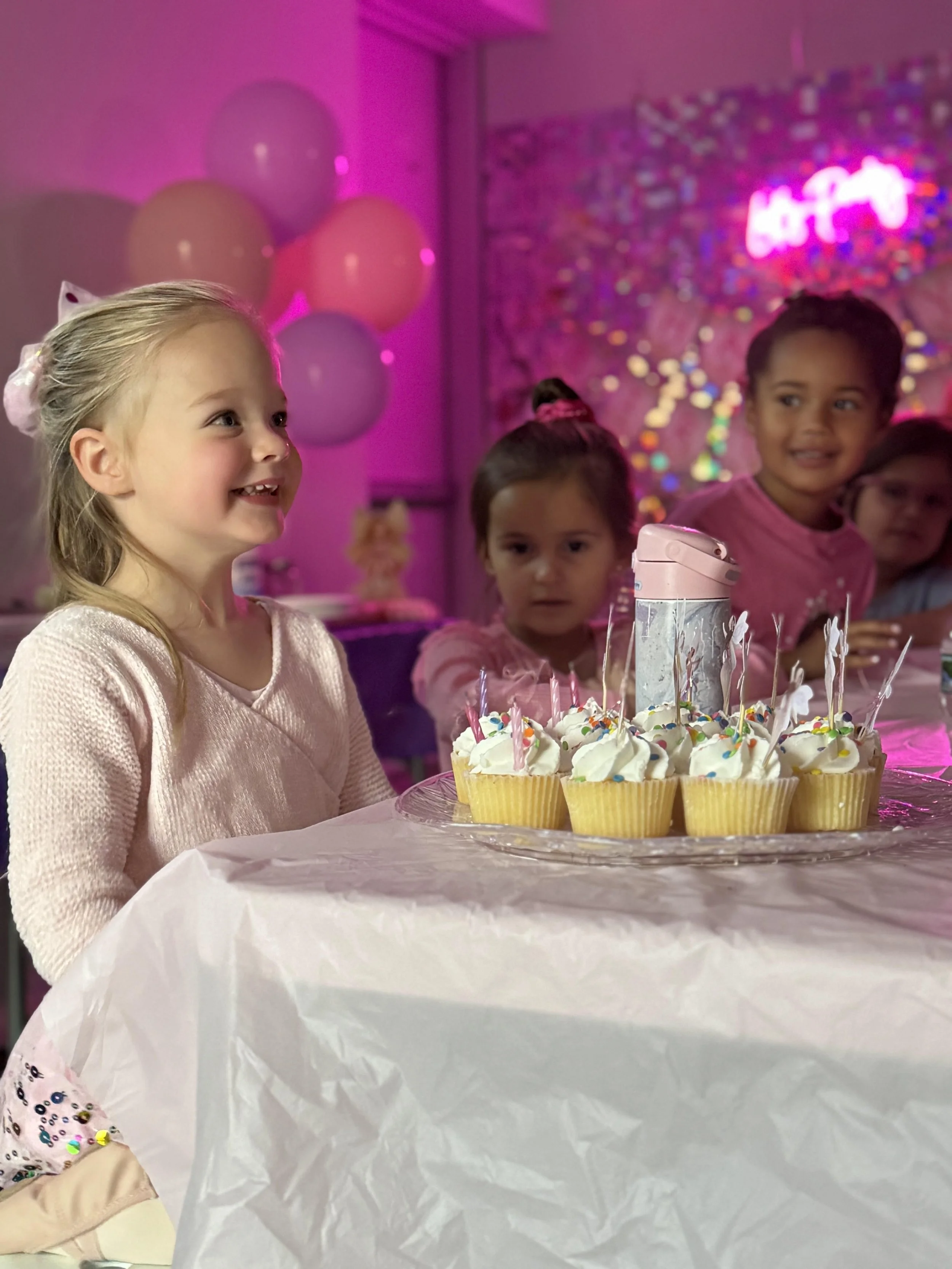 A group of young girls at a birthday party, sitting at a table with cupcakes and candles, decorated with pink balloons and colorful decorations.