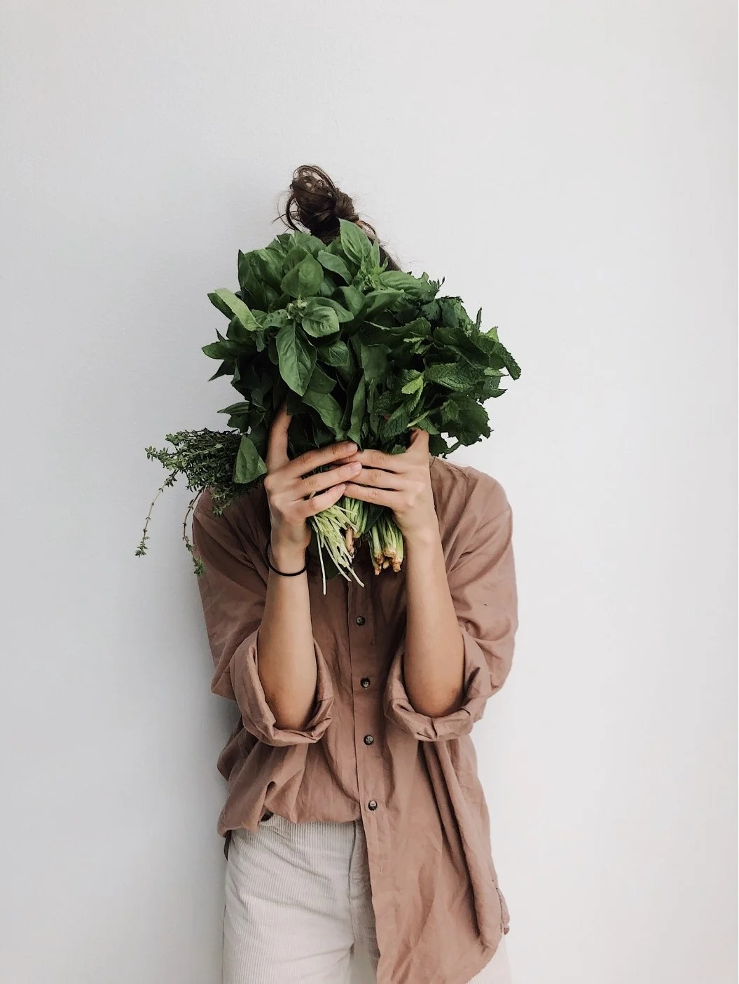 Person holding a large bouquet of green leafy herbs in front of their face, wearing a brown button-up shirt and light-colored pants, standing against a plain white wall.to represent the naturopath carly watkinsherbal medicine at remedy natural health