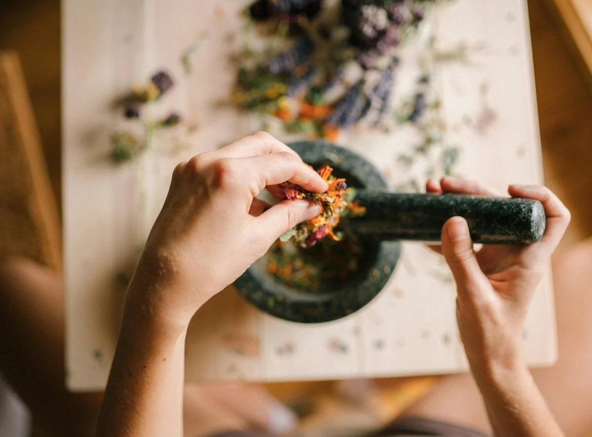 Hands using a pestle and mortar to grind colorful dried herbs and flowers on a wooden table to represent the herbal medicine at remedy natural health
