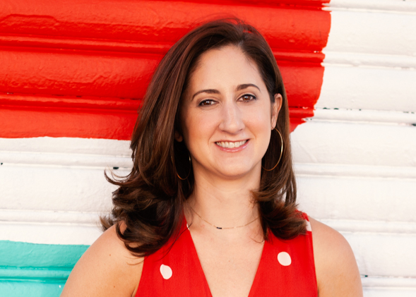 A woman with shoulder-length brown hair, wearing a red sleeveless top with white polka dots, standing in front of a colorful striped background.