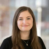 A young woman with shoulder-length brown hair, smiling, wearing a black top and a necklace, standing indoors with blurred background