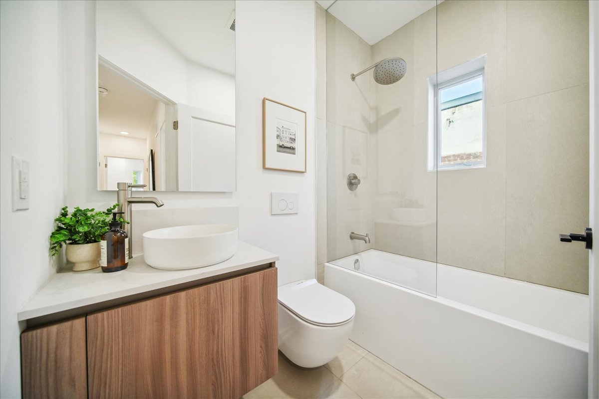 Modern bathroom with white fixtures, floating wood vanity, round sink, large mirror, wall-mounted toilet, shower-tub combo, glass door, and minimal decor.