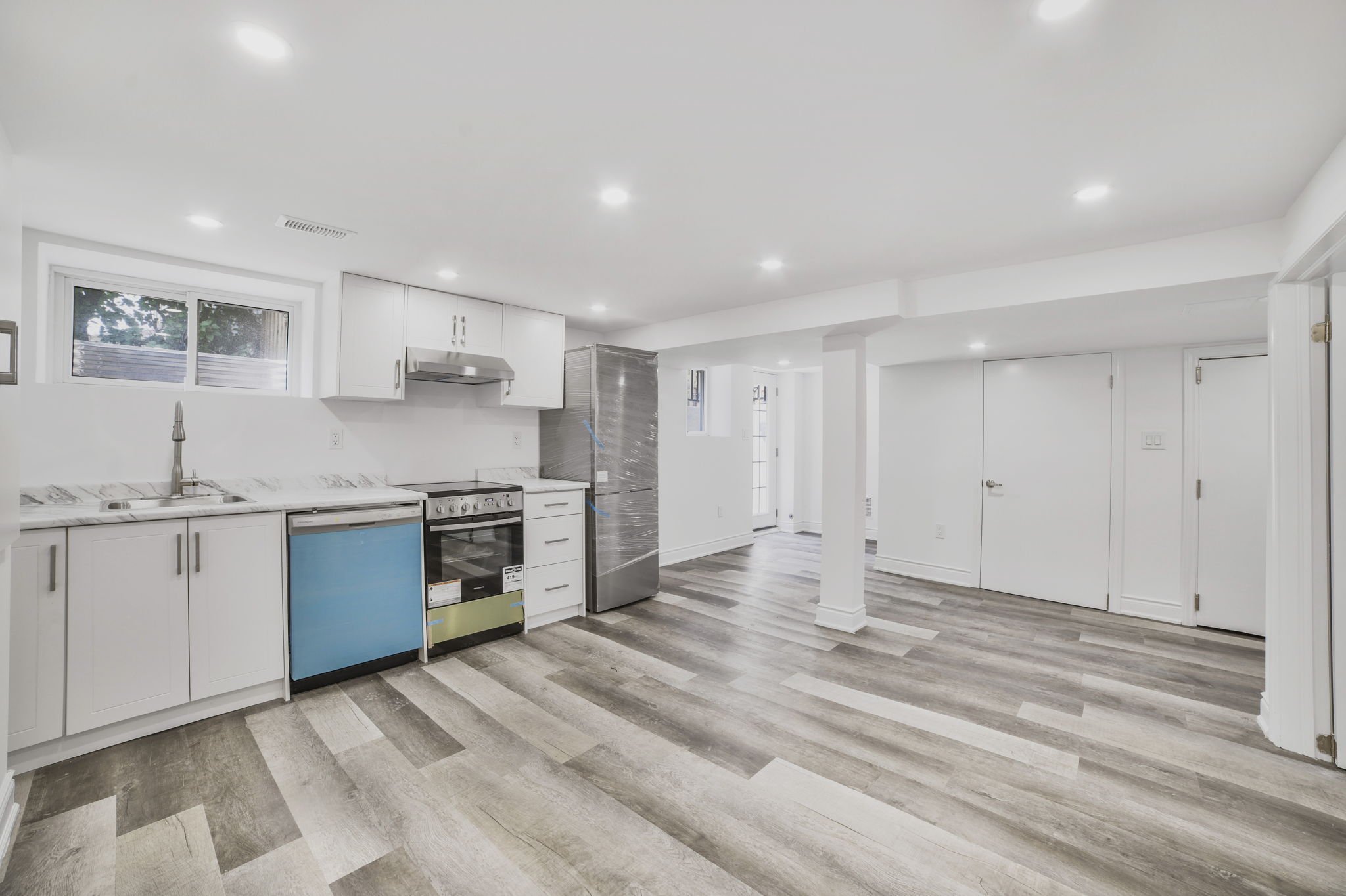 Empty modern kitchen with white cabinets, marble countertops, and stainless steel appliances, including a refrigerator and stove, with a wood-look laminate floor and a window above the sink.