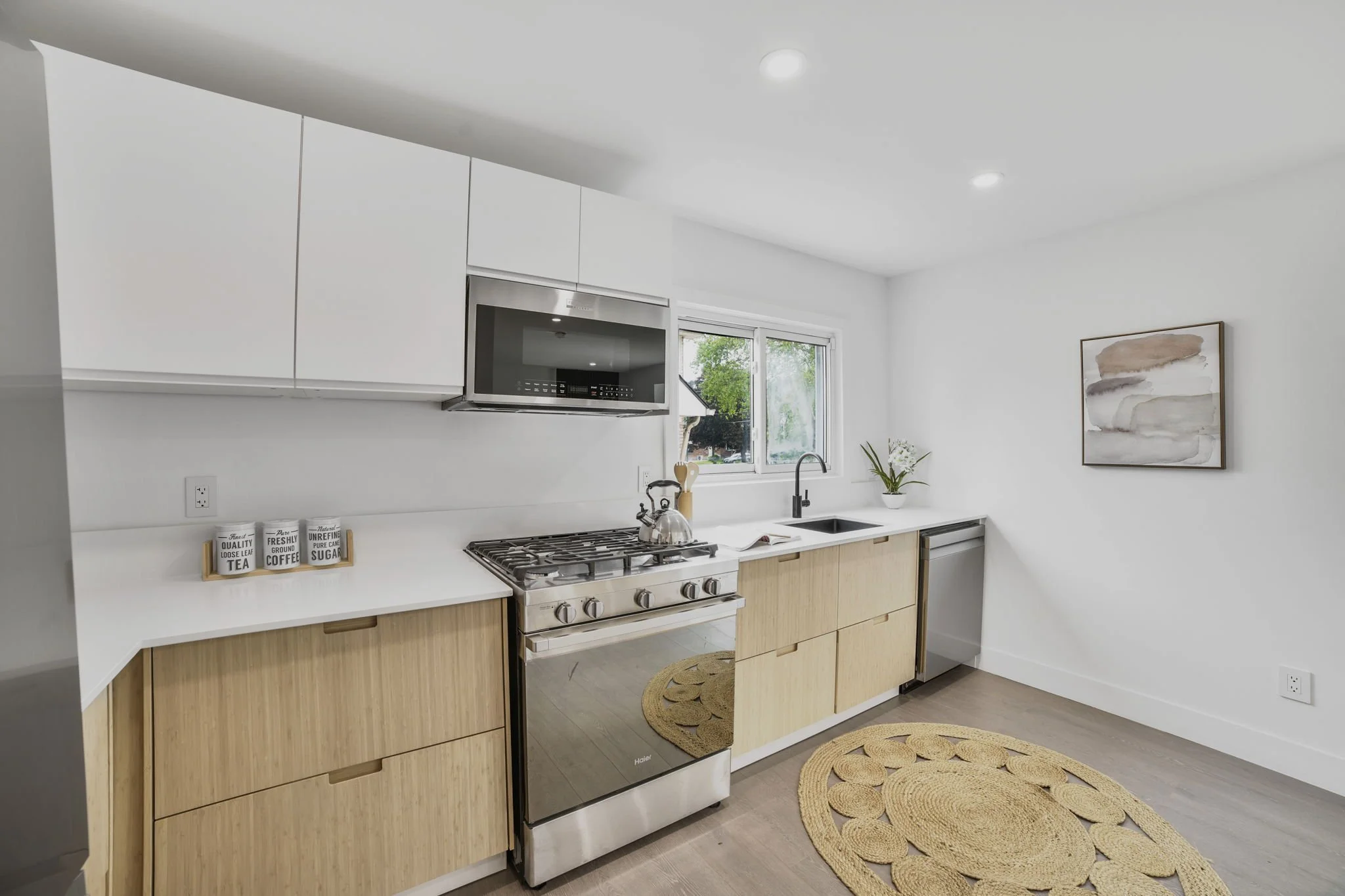 Modern kitchen with white cabinets, a microwave, a stove, a paper towel holder, a window above the sink, and a decorative painting on the wall. Rattan rug on the floor.