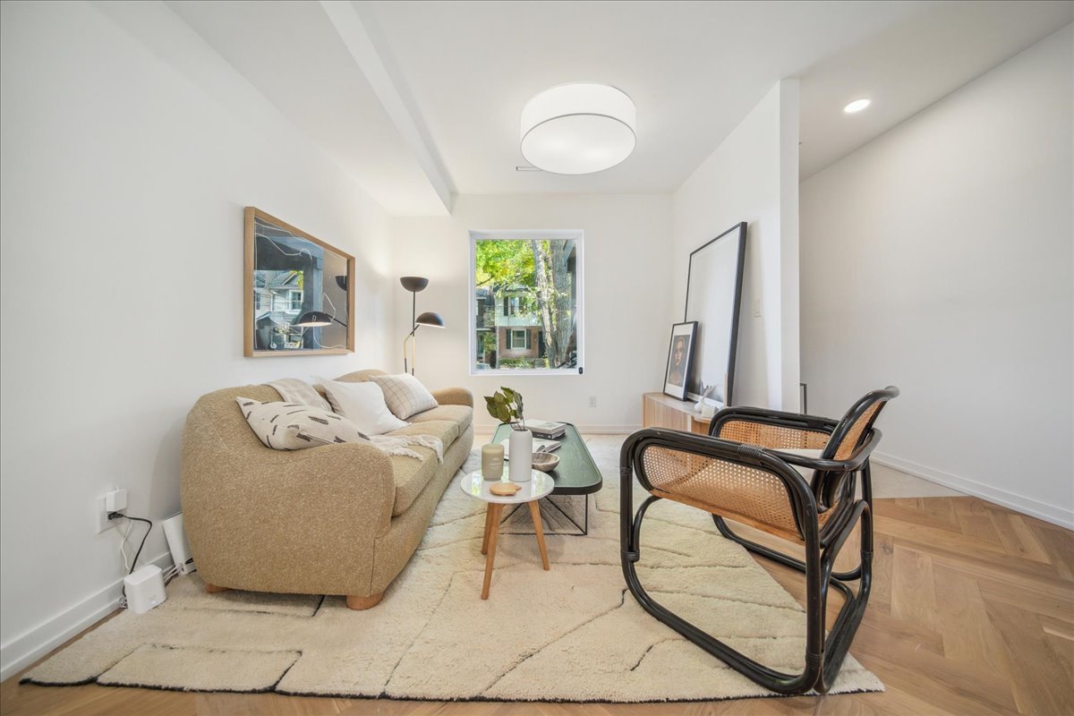 Modern living room with a beige sofa, decorative pillows, a black floor lamp, and a wooden chair. Features a rectangular glass coffee table, framed artwork, indoor plants, and a large window with trees outside.