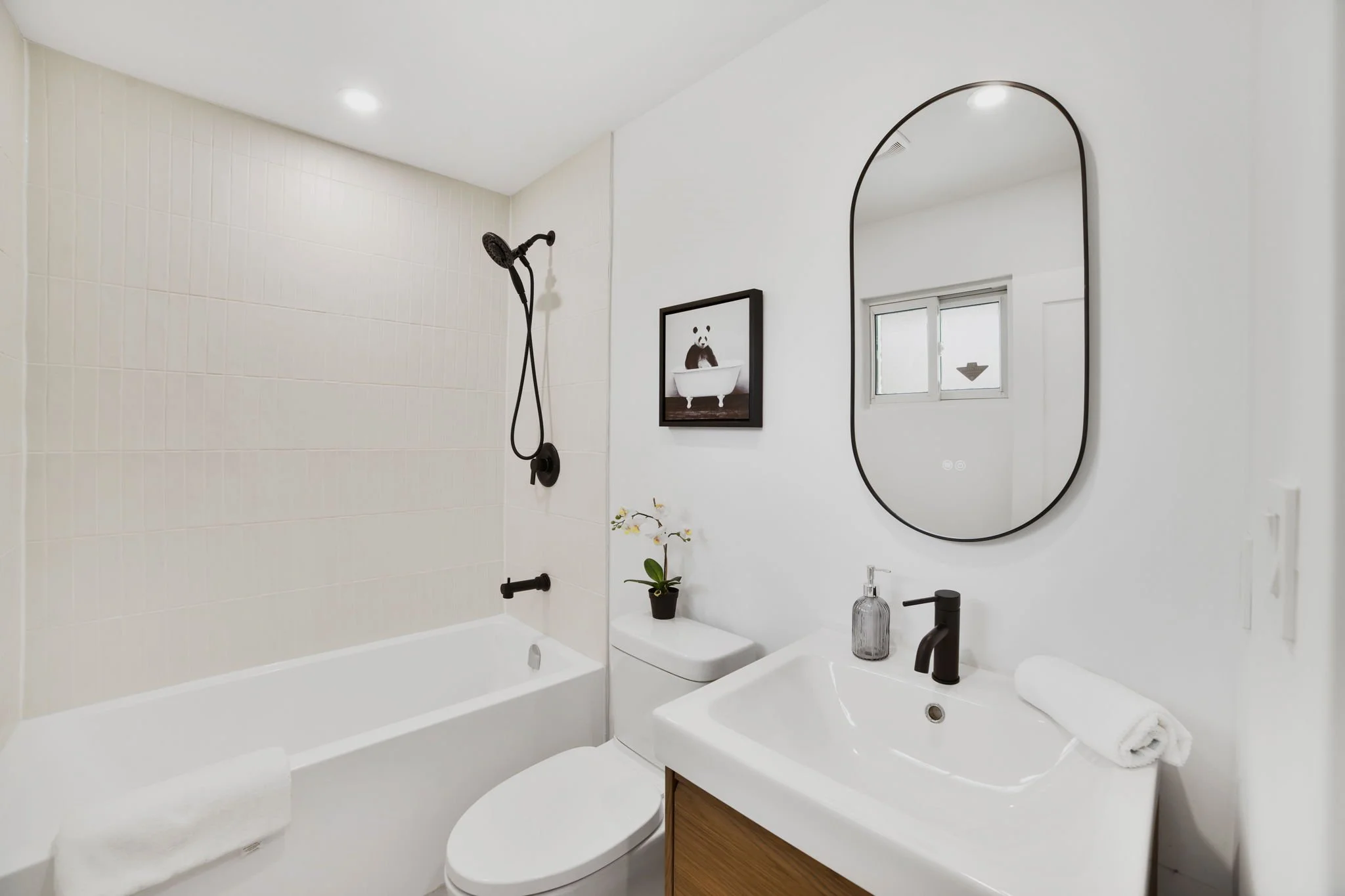 Modern bathroom with white walls, a bathtub and a shower with a black showerhead, a white toilet with a small potted orchid on top, a wooden vanity with a white sink, a black faucet, a rolled white towel, a black soap dispenser, a oval mirror, a fram