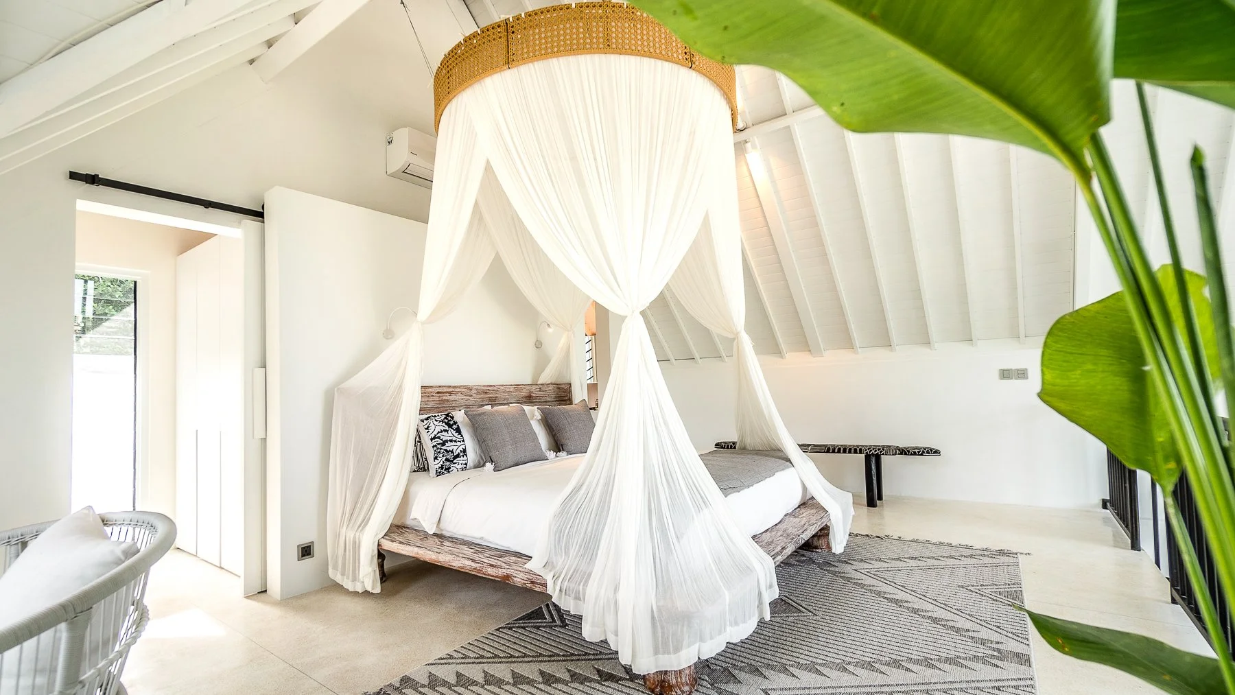 Bedroom with a canopy bed featuring white curtains, a wooden headboard, and gray, black, and white pillows. The room has white walls, a sloped ceiling with white wood panels, and green plants partially visible in the foreground.