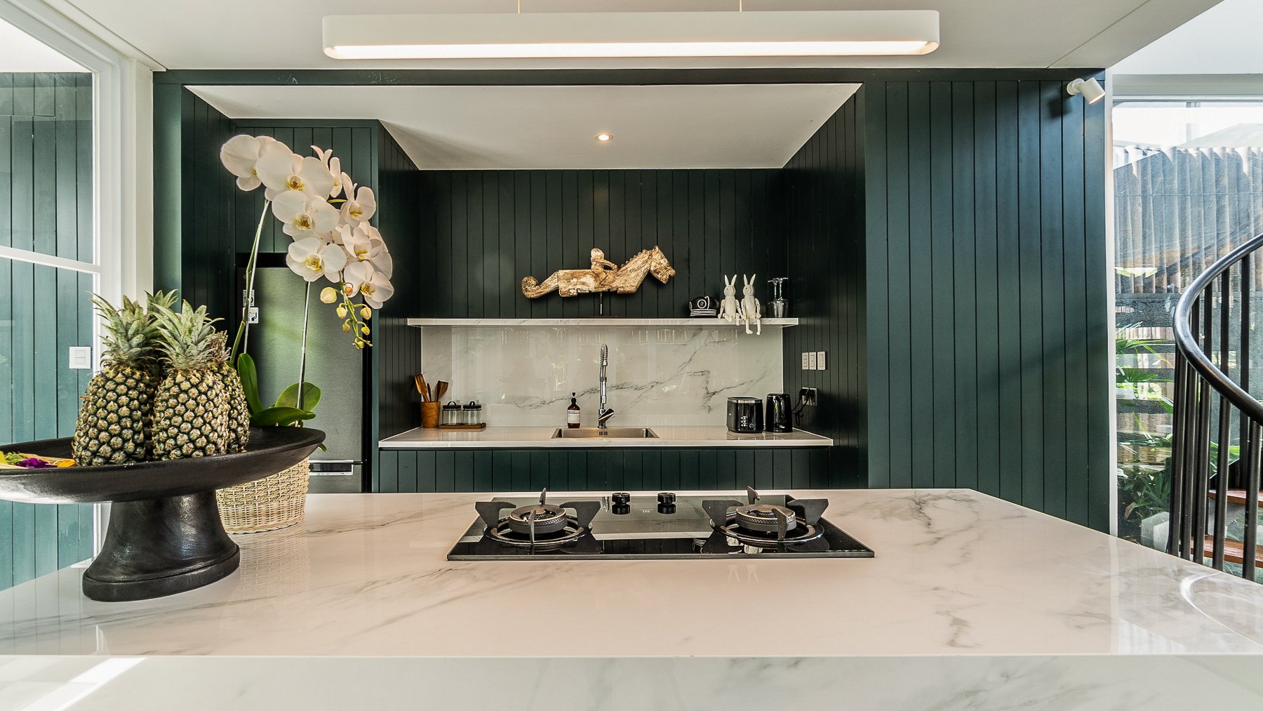 Modern kitchen with marble island countertop, green paneled walls, and decorative items, including a bowl of pineapples and a potted orchid plant, with a stove built into the counter and various small appliances and decor on the back shelf.