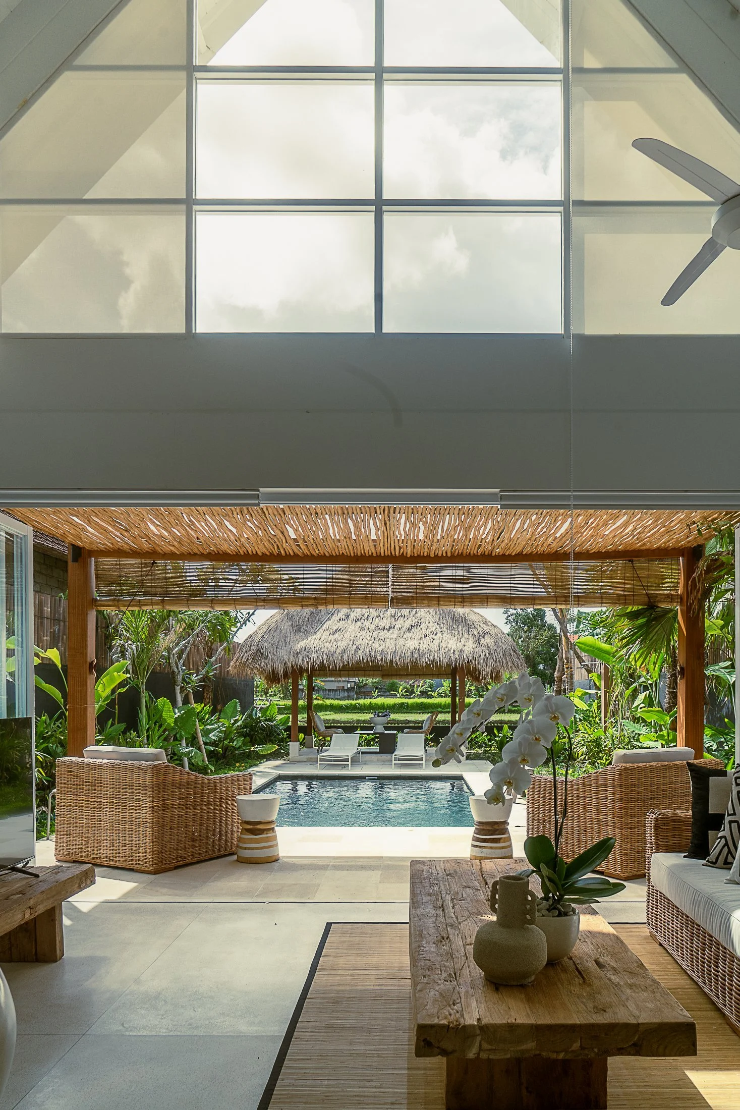 Interior view of a tropical-style home with an open patio overlooking a pool and lush garden, featuring wicker furniture, potted plants, and a thatched roof gazebo.