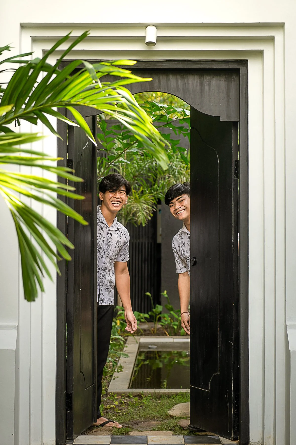 Two young men peeking through a black wooden gate, smiling and laughing, surrounded by plants and greenery.