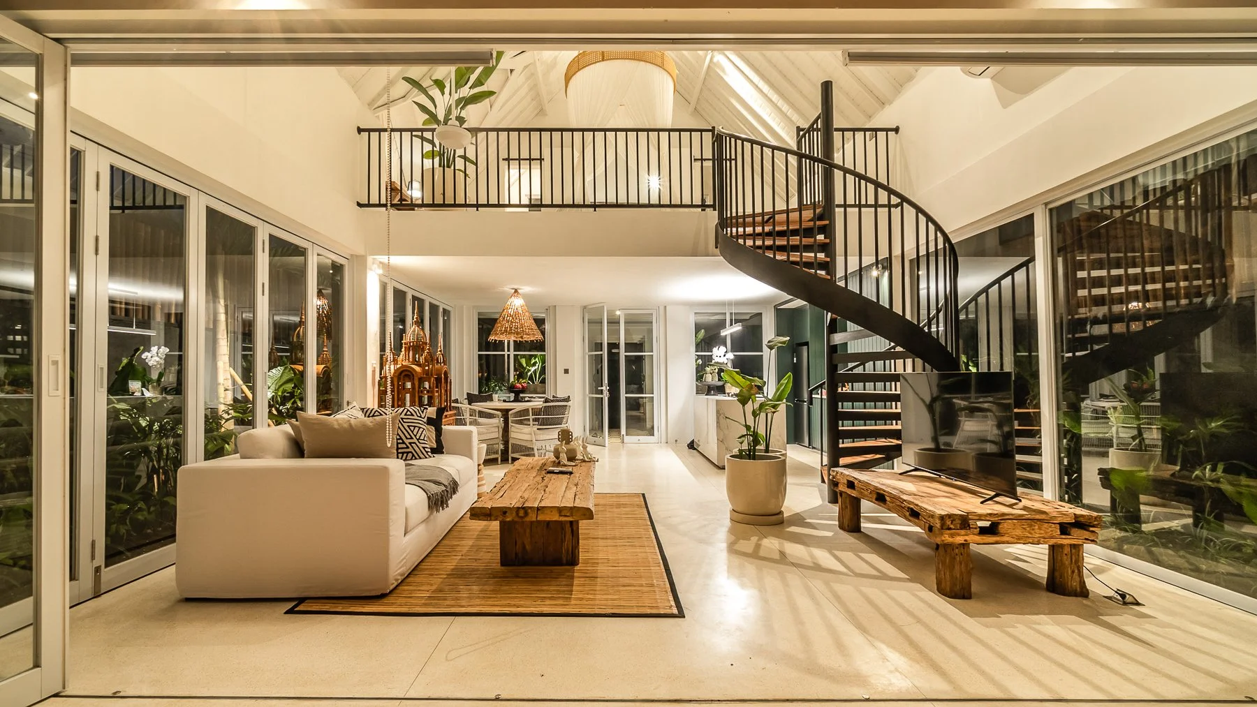Open concept living room with white sofa, wooden coffee table, green plants, glass sliding doors, and a black spiral staircase leading to a second floor.