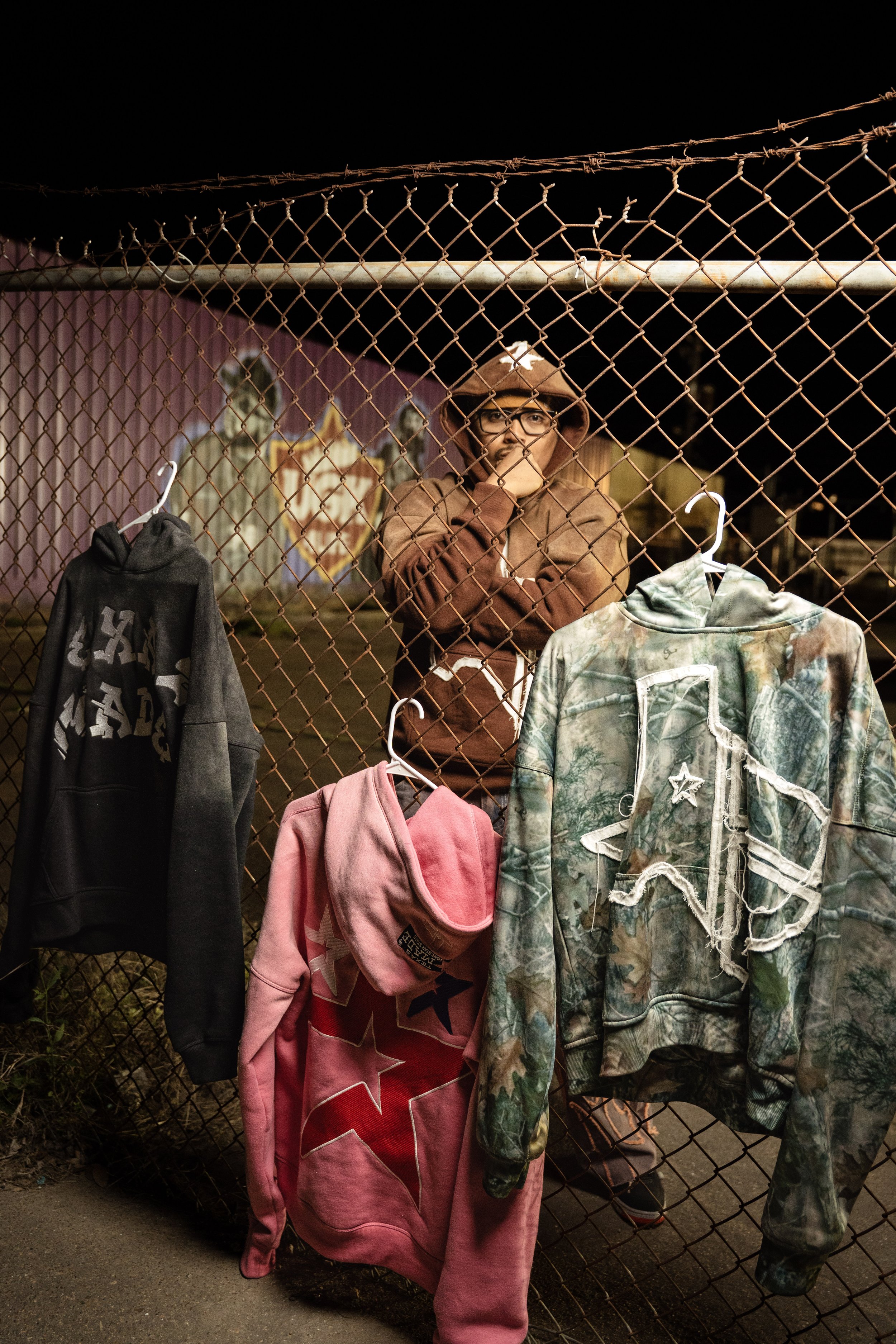 A person stands behind a chain-link fence at night, wearing a brown hoodie with a logo. Three hoodies hang on the fence: a black one, a pink one with stars, and a camo-patterned one with a Texas outline.